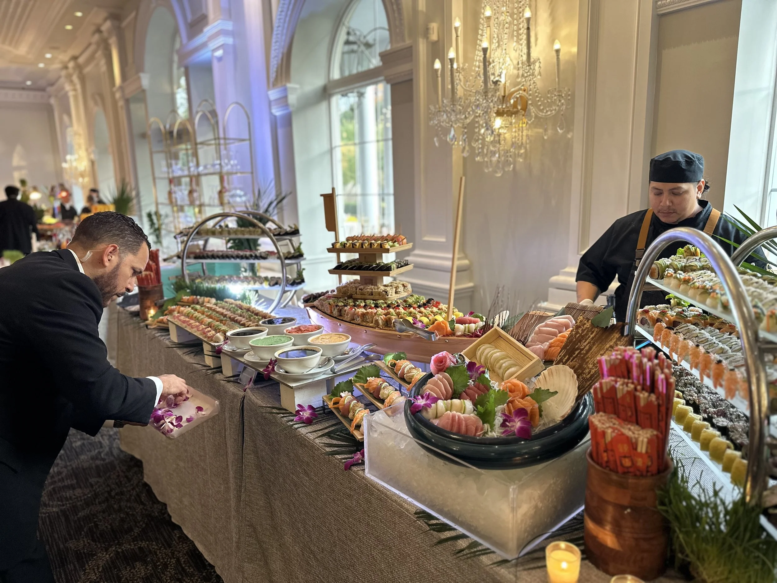 Buffet table with an array of sushi and sashimi, decorative flowers, sauces, and desserts, in a luxurious event space with chandeliers and elegant decor.