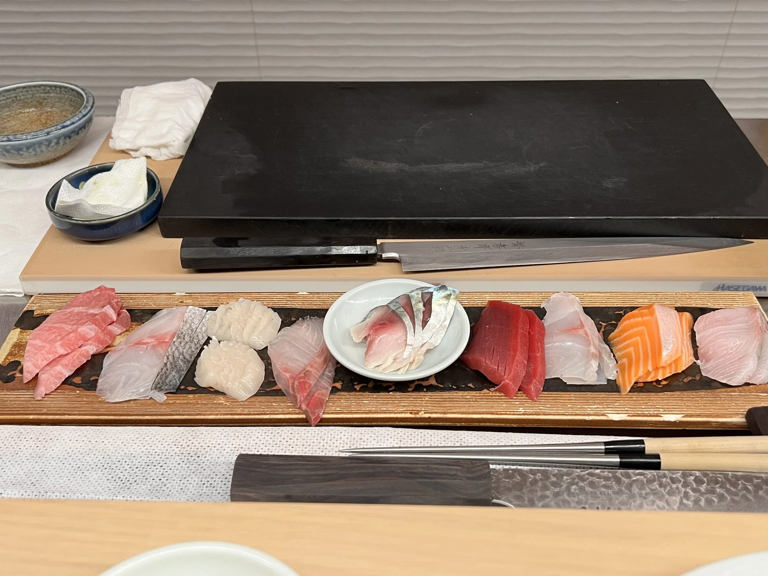 A sushi platter with various types of raw fish such as salmon, tuna, and yellowtail, presented on a wooden board. There is a small bowl of sliced fish, a black cutting board with a knife, and two bowls of sauce and napkins in the background.