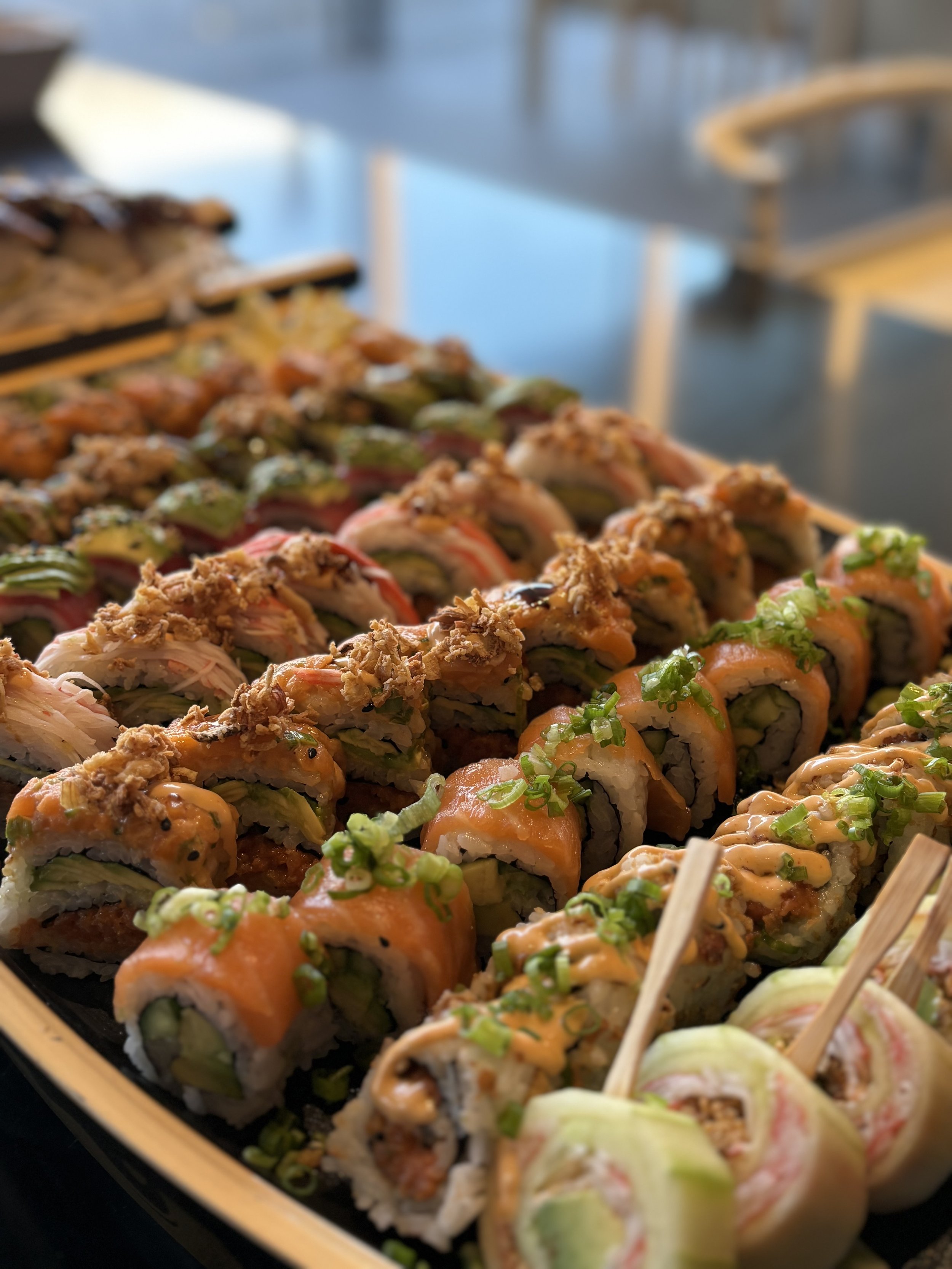 Assorted sushi rolls with different fillings and toppings, arranged in a black container, garnished with green onions and sauces.