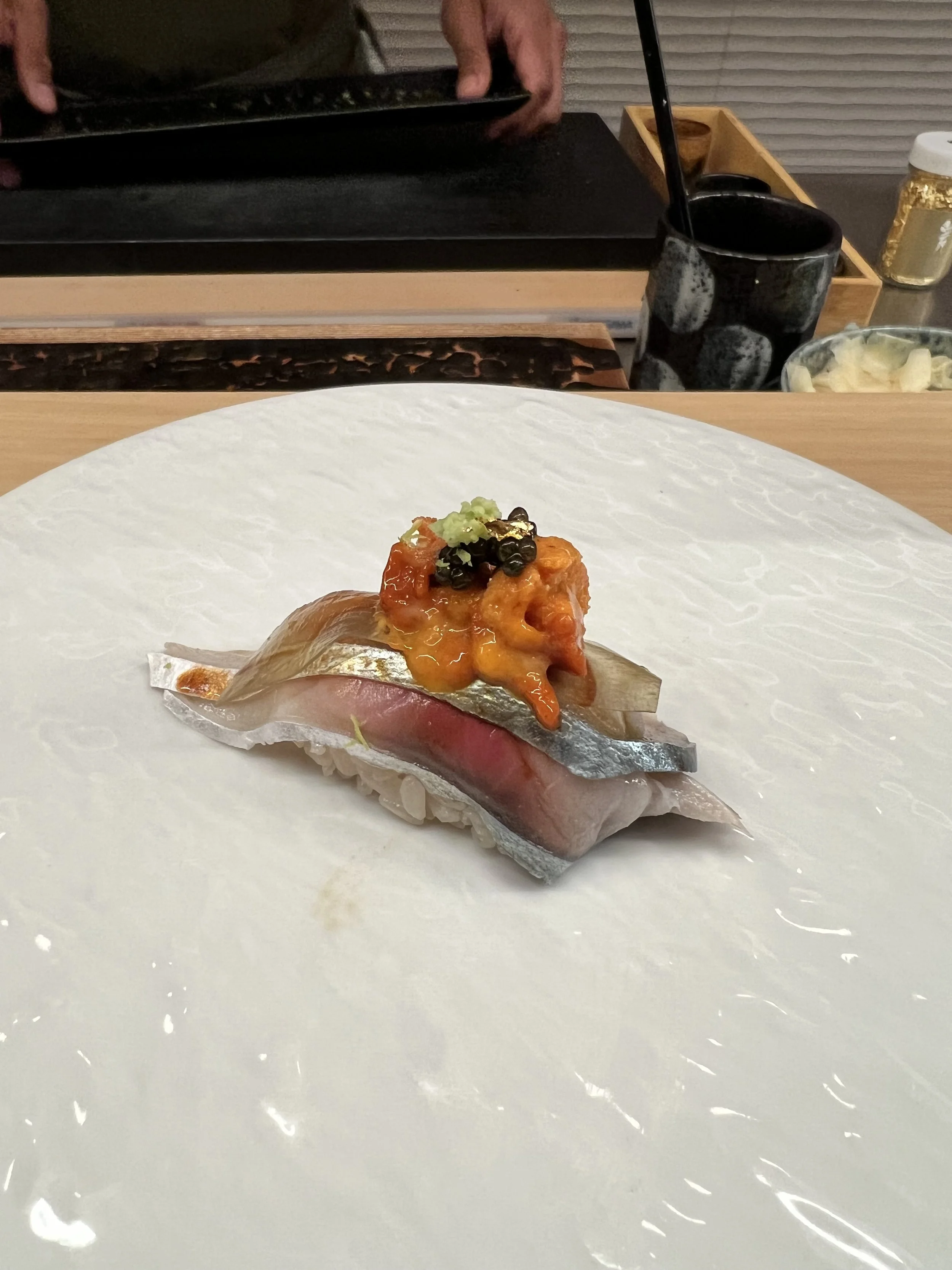 A piece of sushi topped with orange sauce, black beans, and green wasabi on a white paper plate.