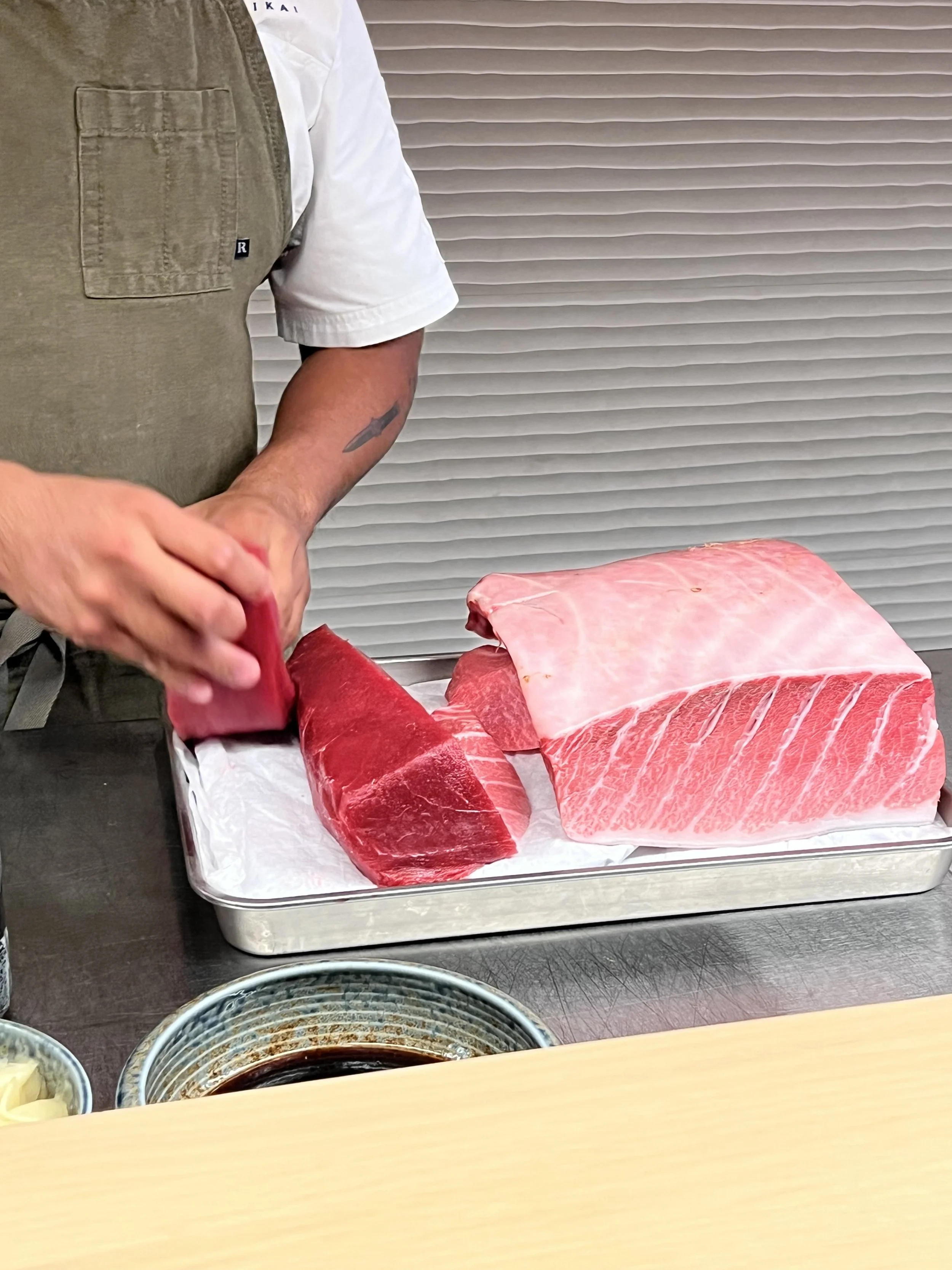 Various cuts of raw beef on a metal tray, with small bowls of dipping sauces and condiments in the foreground, and a person wearing beige pants and a white shirt preparing the meat.