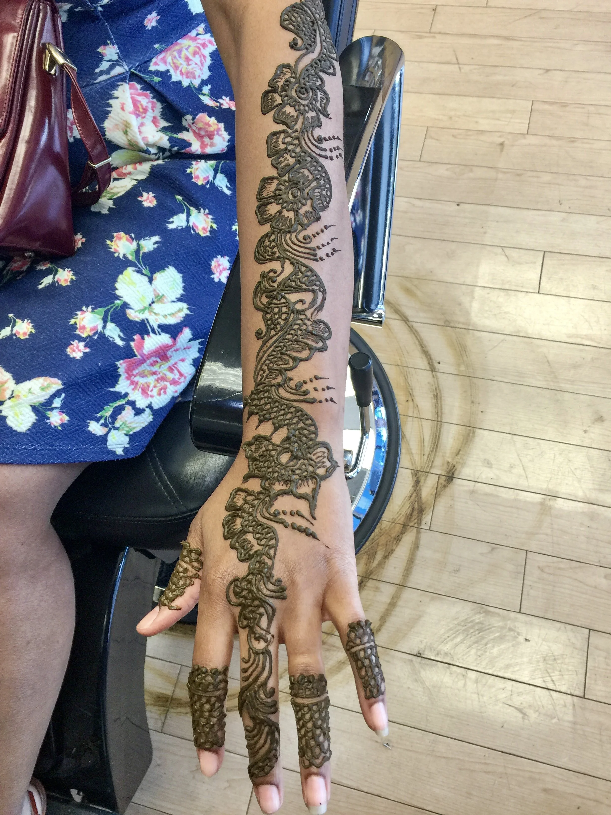 A person's arm and hand with intricate henna-style tattoo designs, featuring a dragon and floral patterns, with a floral dress and a maroon handbag nearby.