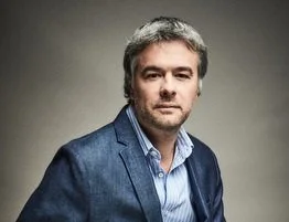 Hombre con barba y cabello gris, vestido con saco azul y camisa, fondo gris