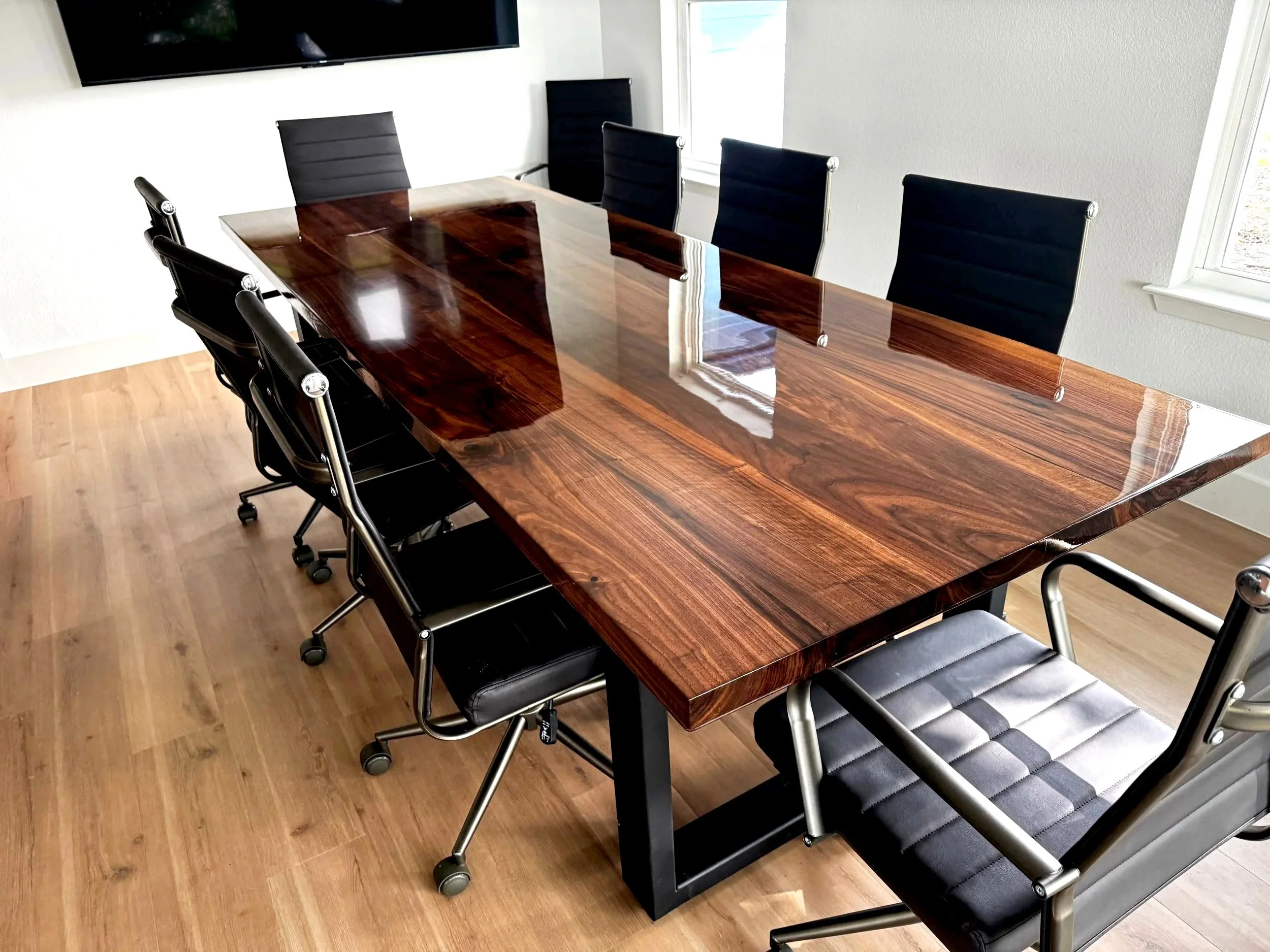 A conference room with a large wooden table and eight black office chairs, four around each side. A flat screen TV is mounted on the wall and a window on the right side of the room letting in natural light.
