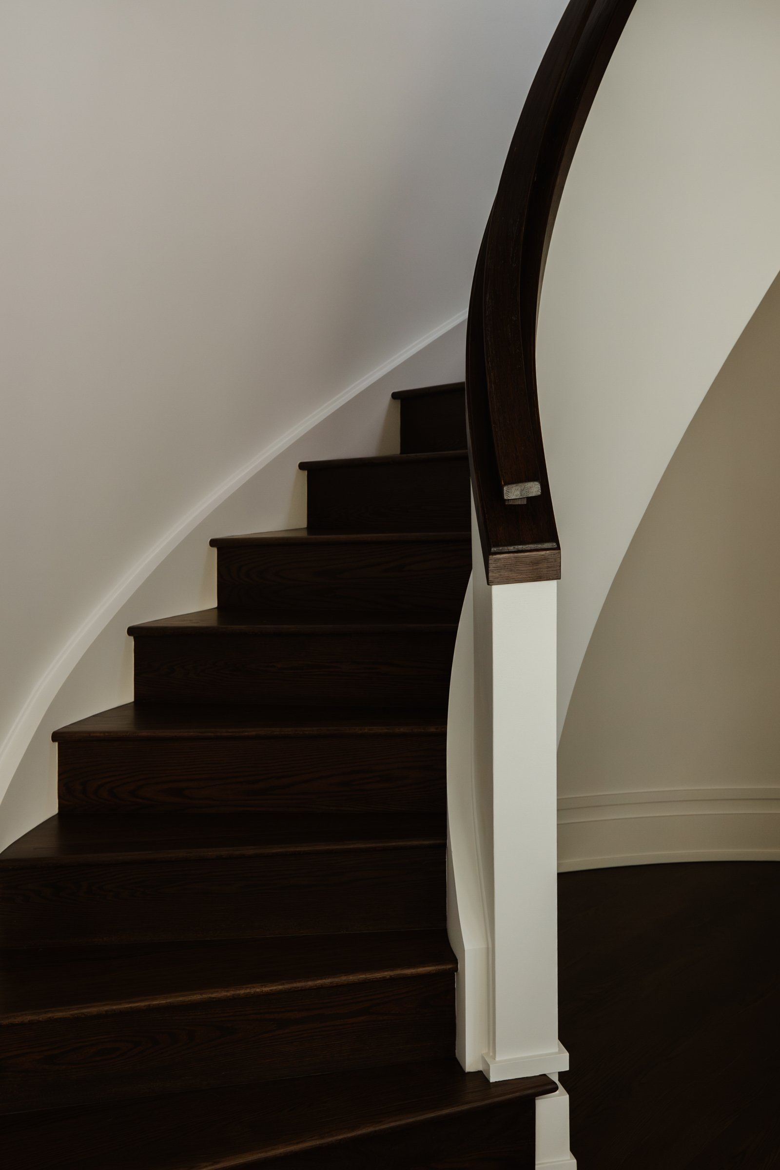 Curved wooden staircase with dark polished steps and a smooth handrail, set against cream-colored wall.