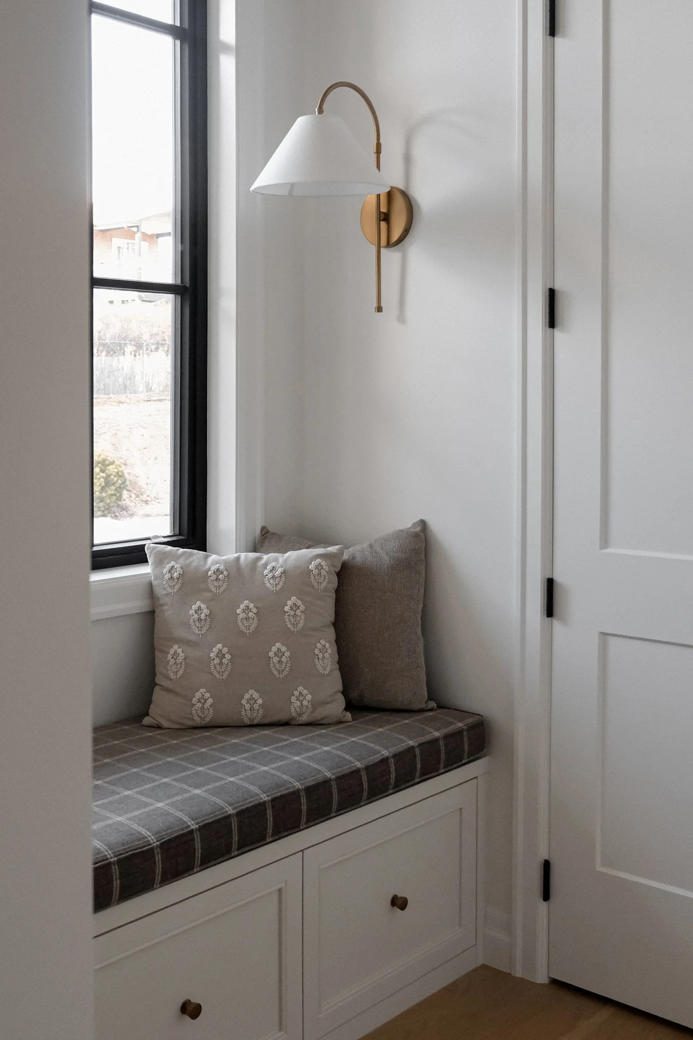 Cozy nook with plaid cushions, featuring two pillows - one with floral embroidery and the other plain. Set against a light wall with a closed door.