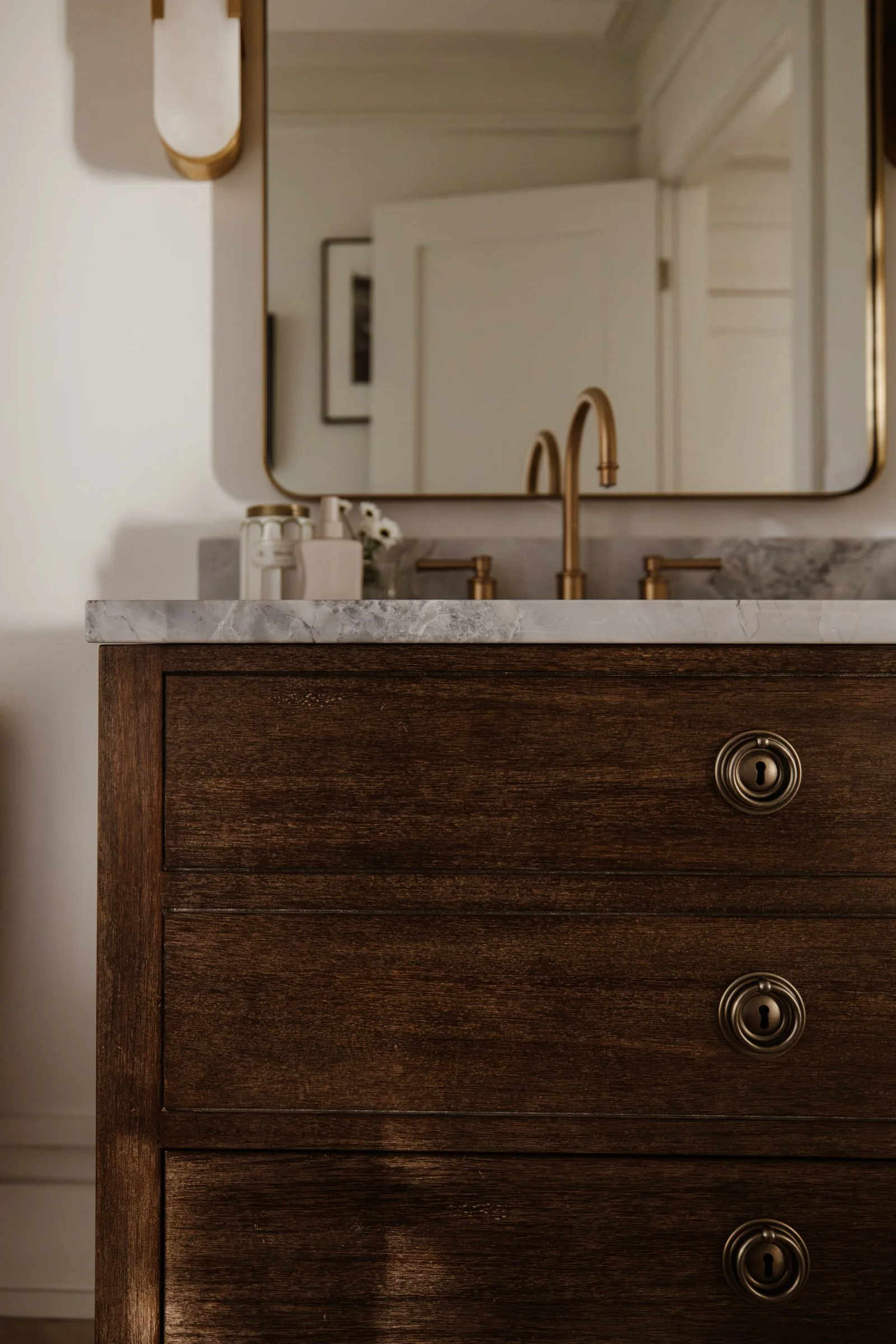 Close-up of a wooden bathroom vanity with round silver knobs. A marble countertop and brass faucet are above, with toiletries and flowers beside it.