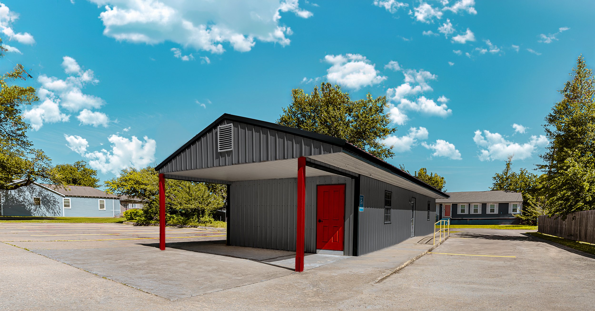 Small gray building with a red door and red support beams, located in a parking lot, with trees and residential buildings and a bright blue sky with clouds in the background.