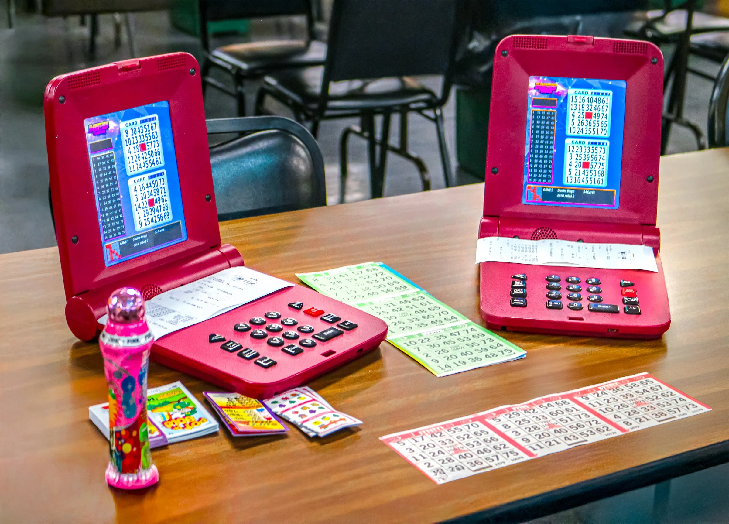 Two pink electronic bingo machines on a wooden table, surrounded by bingo cards, small toys, and a spray bottle, with chairs in the background.