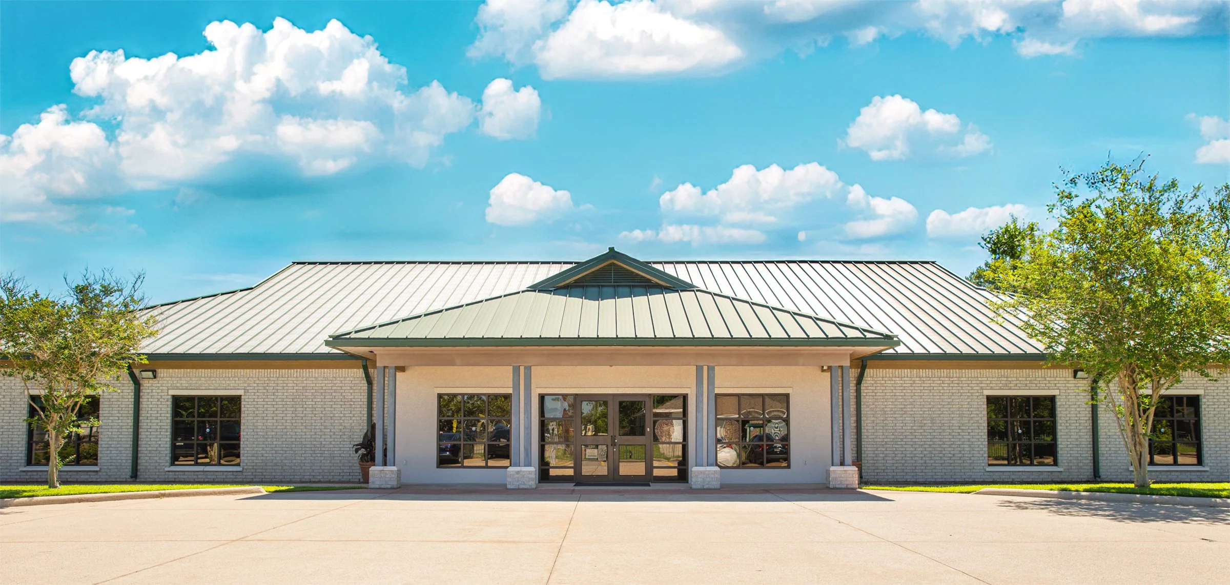 A single-story building with a metal roof, brick exterior, large windows, and a small porch with columns, surrounded by trees under a partly cloudy sky.