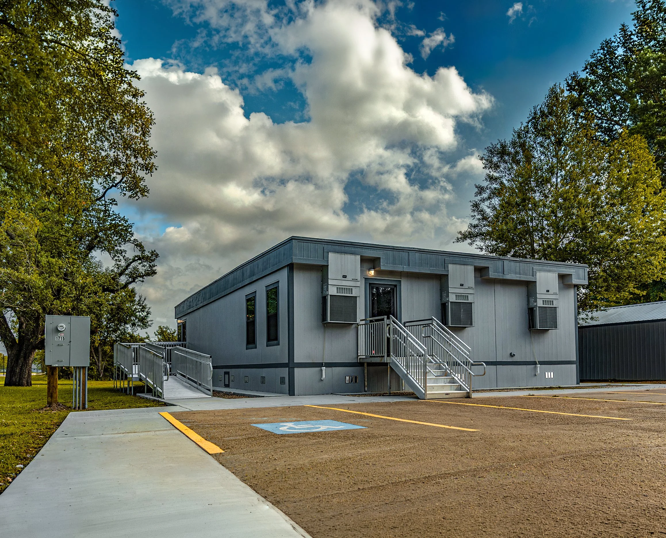A small building with stairs leading up to an entrance surrounded by trees and a parking lot with a handicap space marked on the ground, under a partly cloudy sky.