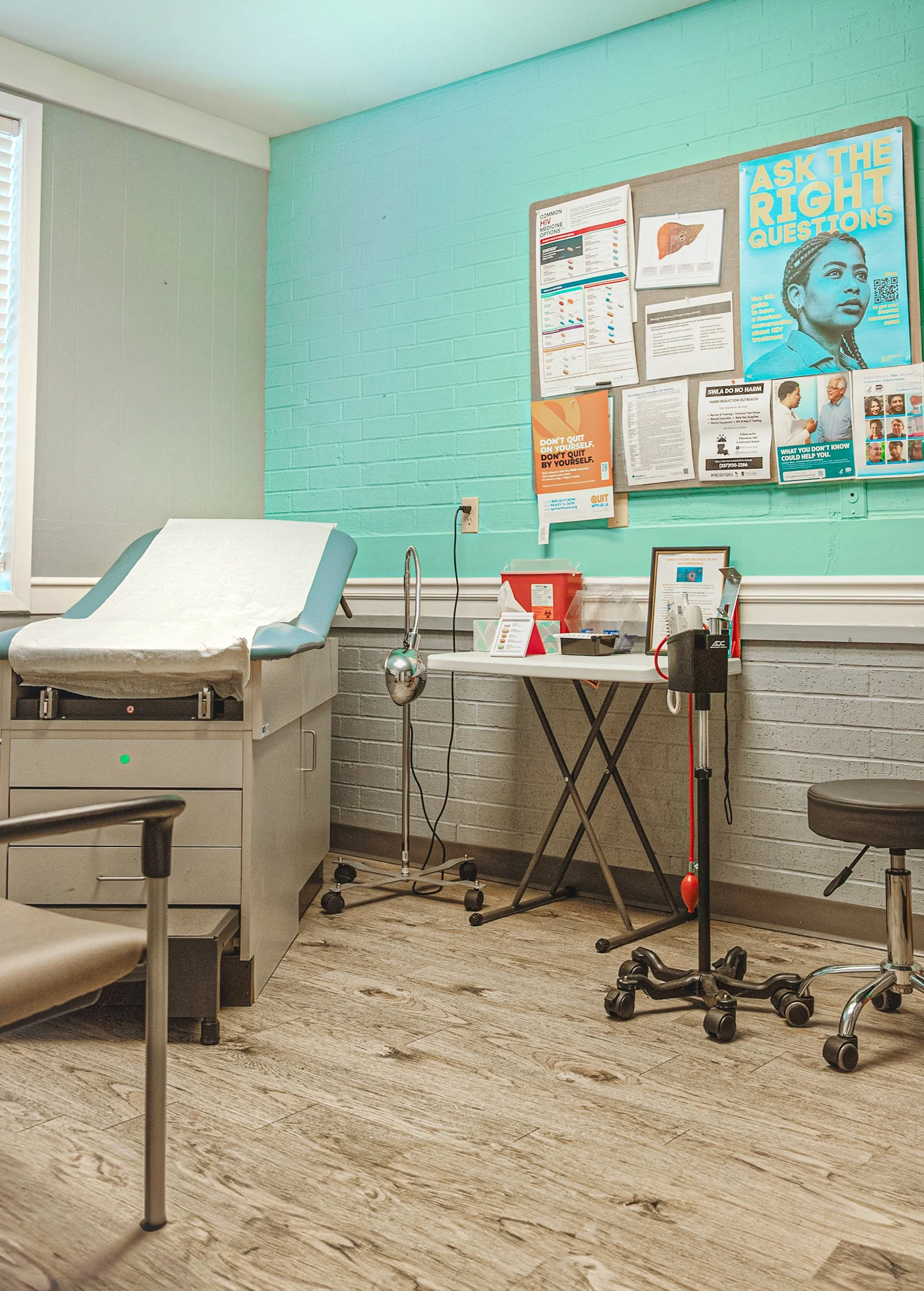 A medical examination room with a hospital bed covered with a white sheet, a rolling IV stand, a table with medical supplies, and a bulletin board on a greenish wall displaying informational posters and notices. The room has wood-like flooring and appears clean and organized.
