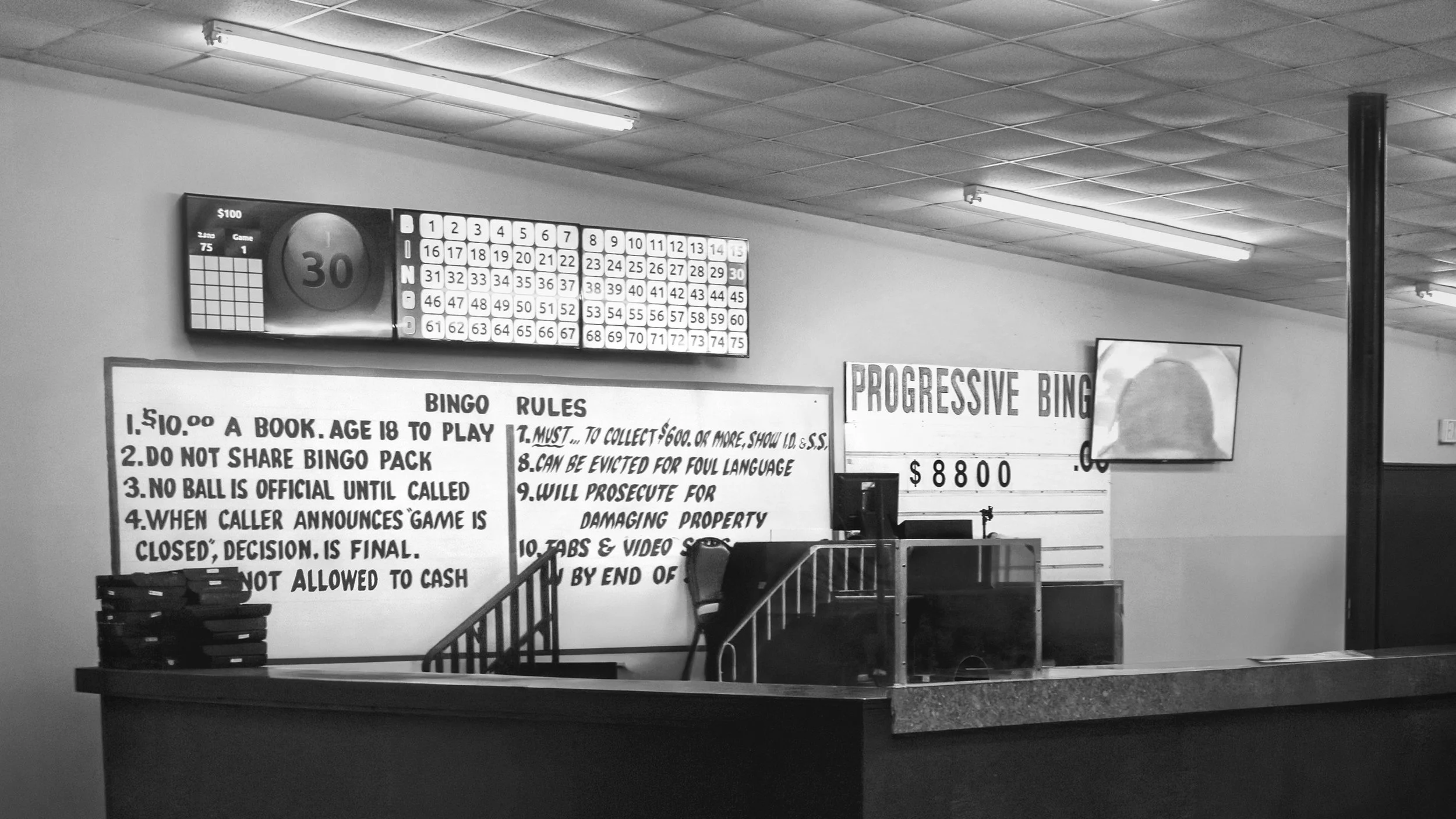 An indoor bingo hall with digital displays and rules. The display shows a bingo game with numbers and a timer. There's a sign with bingo rules and a progressive bingo payout of $8800. The room has fluorescent ceiling lights and a counter with stacked trays.
