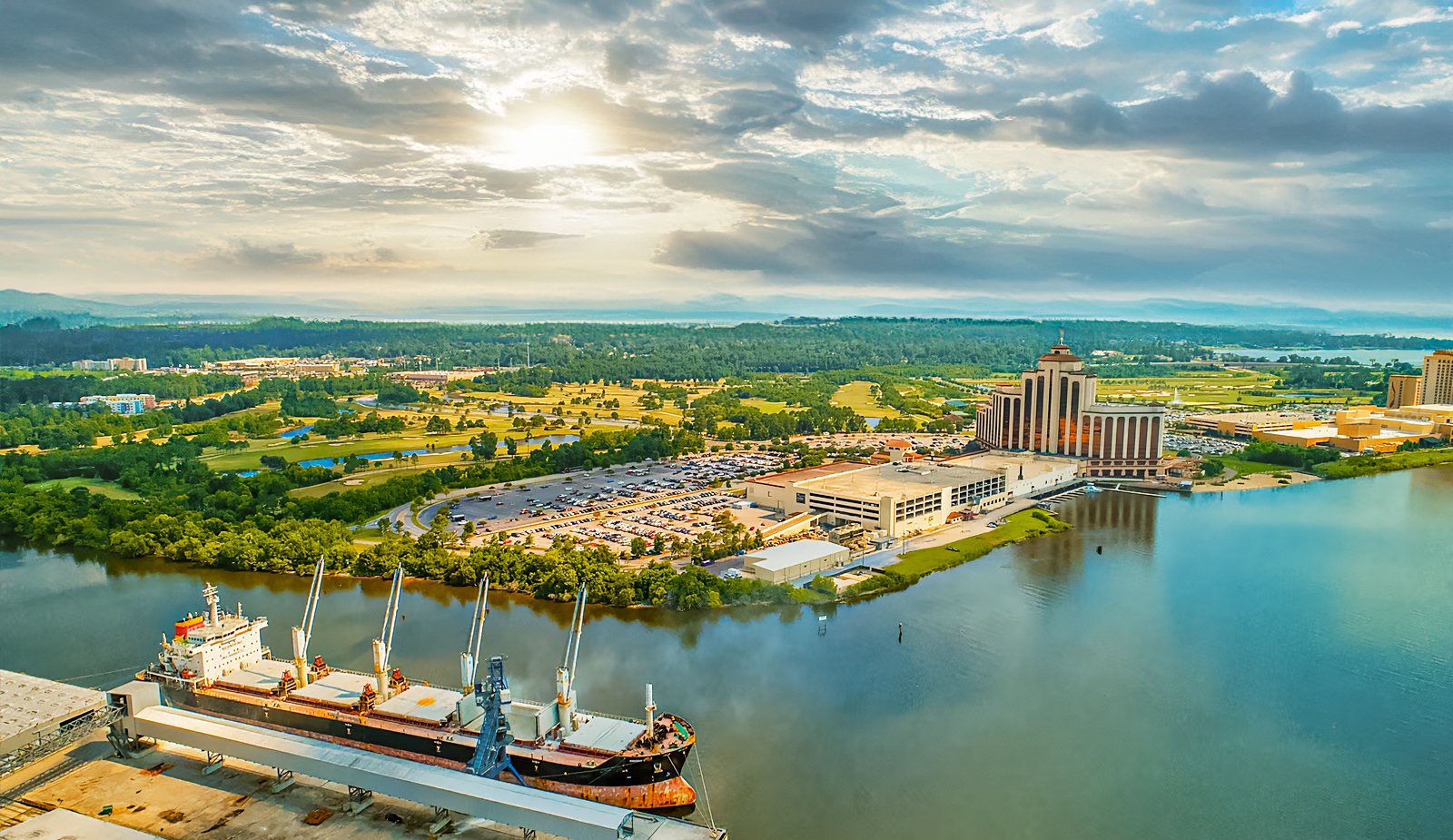 Aerial view of a city featuring a large river with a docked ship, tall buildings including a prominent multi-story structure, and a vast green landscape with fields and forests under a partly cloudy sky at sunset.