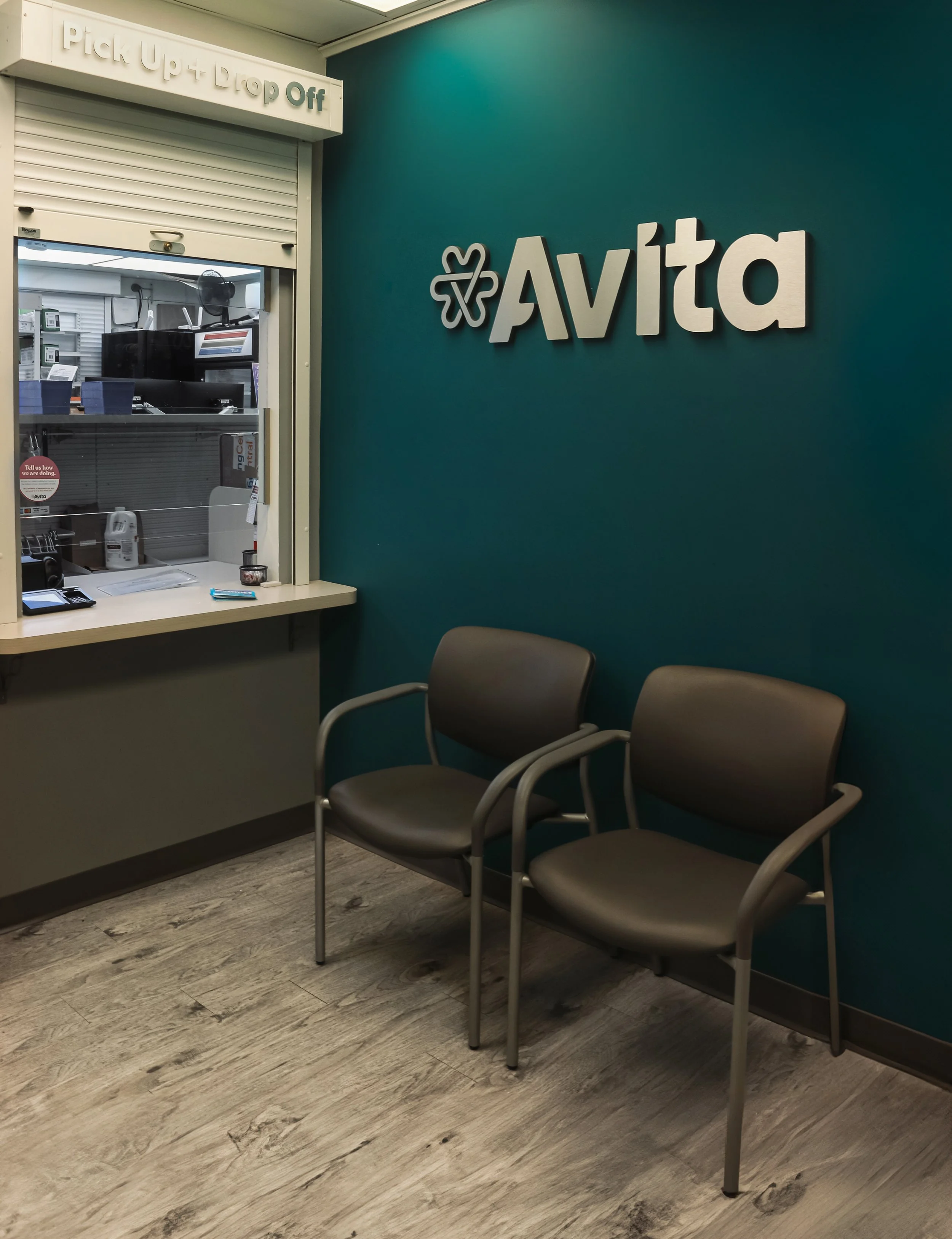 Waiting room with two chairs in front of teal wall with 'Avita' sign; reception window with office supplies, medical equipment, and a computer monitor visible behind glass.