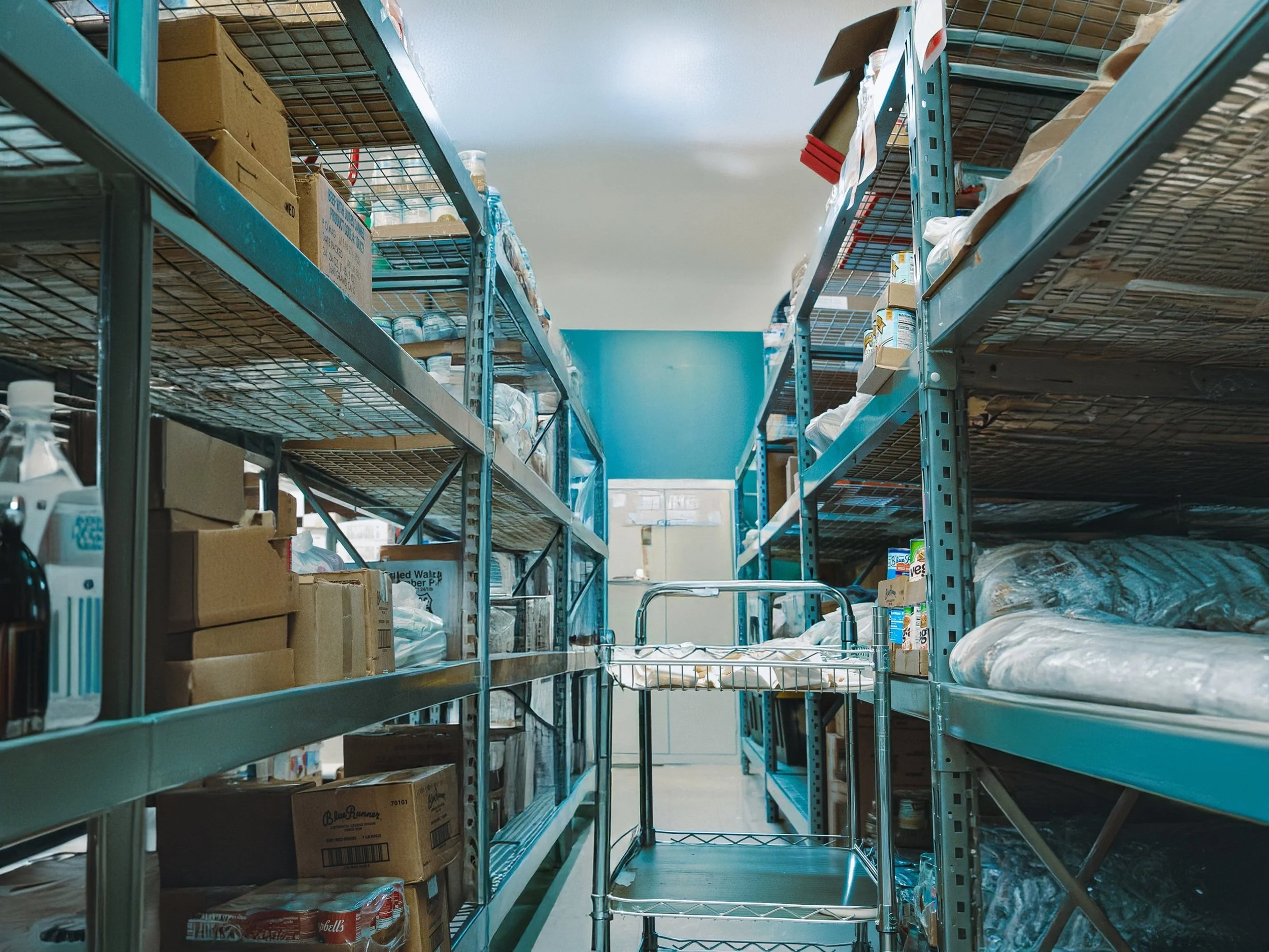 Metal storage shelves in a warehouse filled with cardboard boxes, canned goods, and other packaged products.