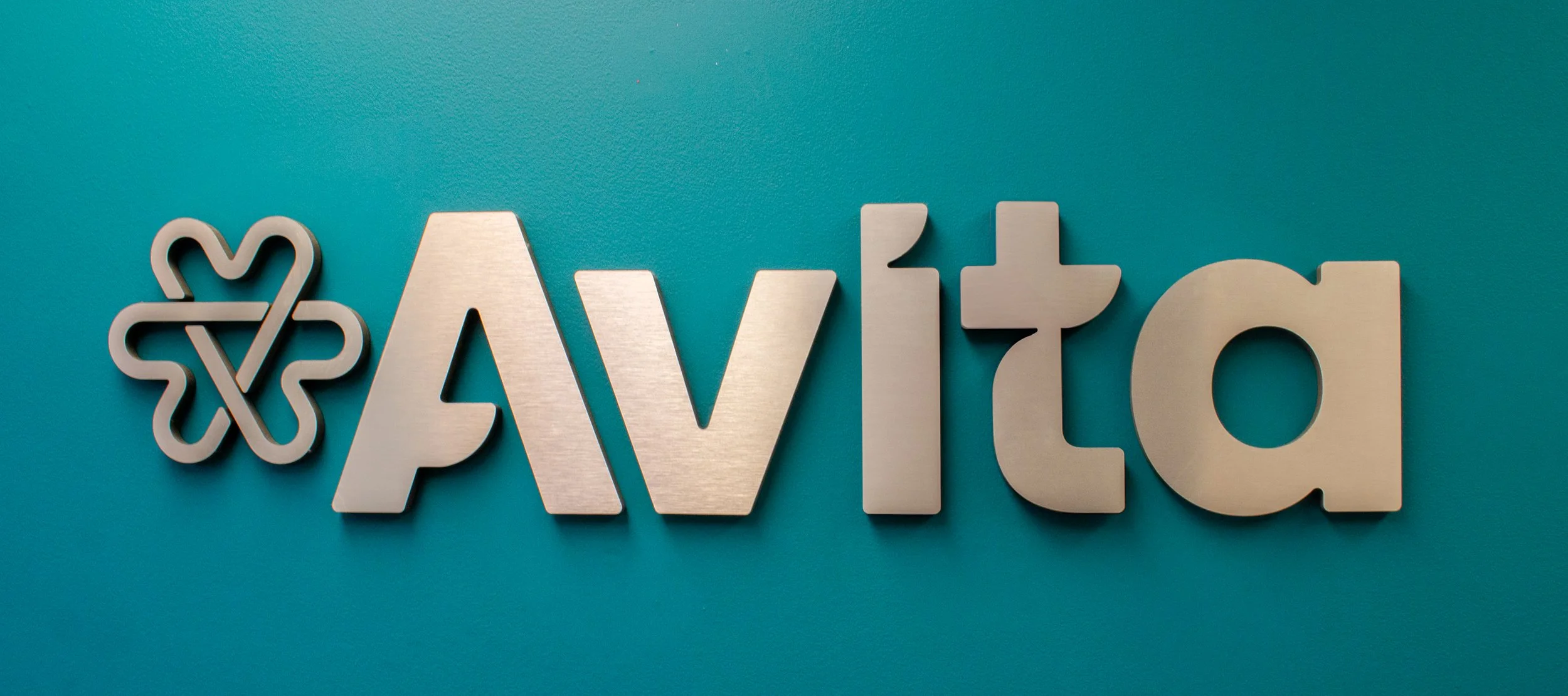 Metal sign spelling out 'Avito' with a four-leaf clover graphic on a teal wall.