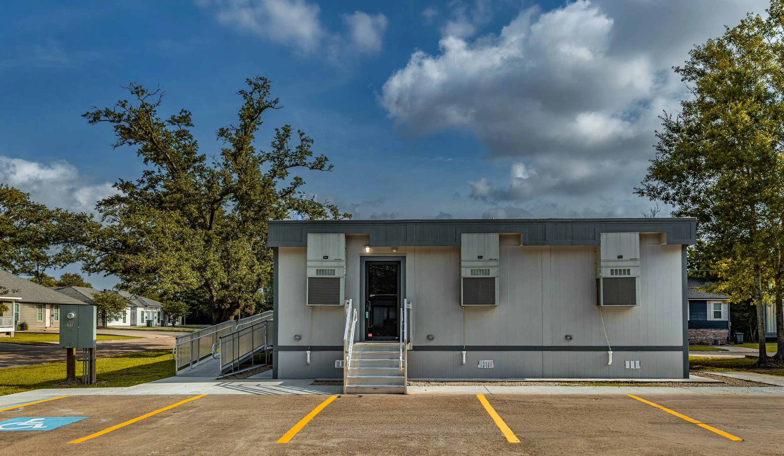 A small portable building with stairs and windows, surrounded by a parking lot with marked parking spaces, including a handicapped space, and trees in the background, under a partly cloudy sky.