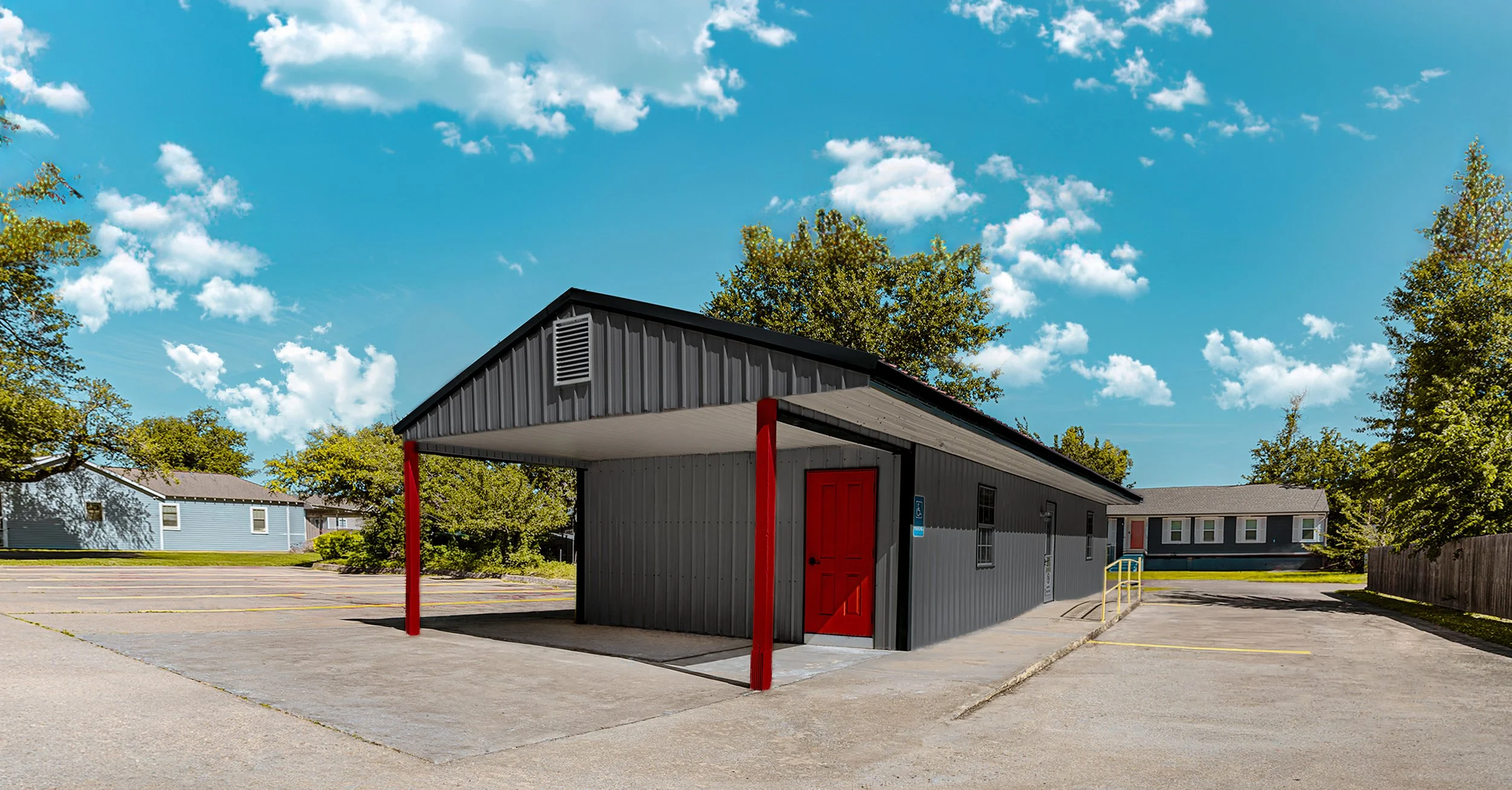 A small gray building with a red door and red support beams, situated in a parking lot with another light blue building and green trees in the background, under a partly cloudy blue sky.