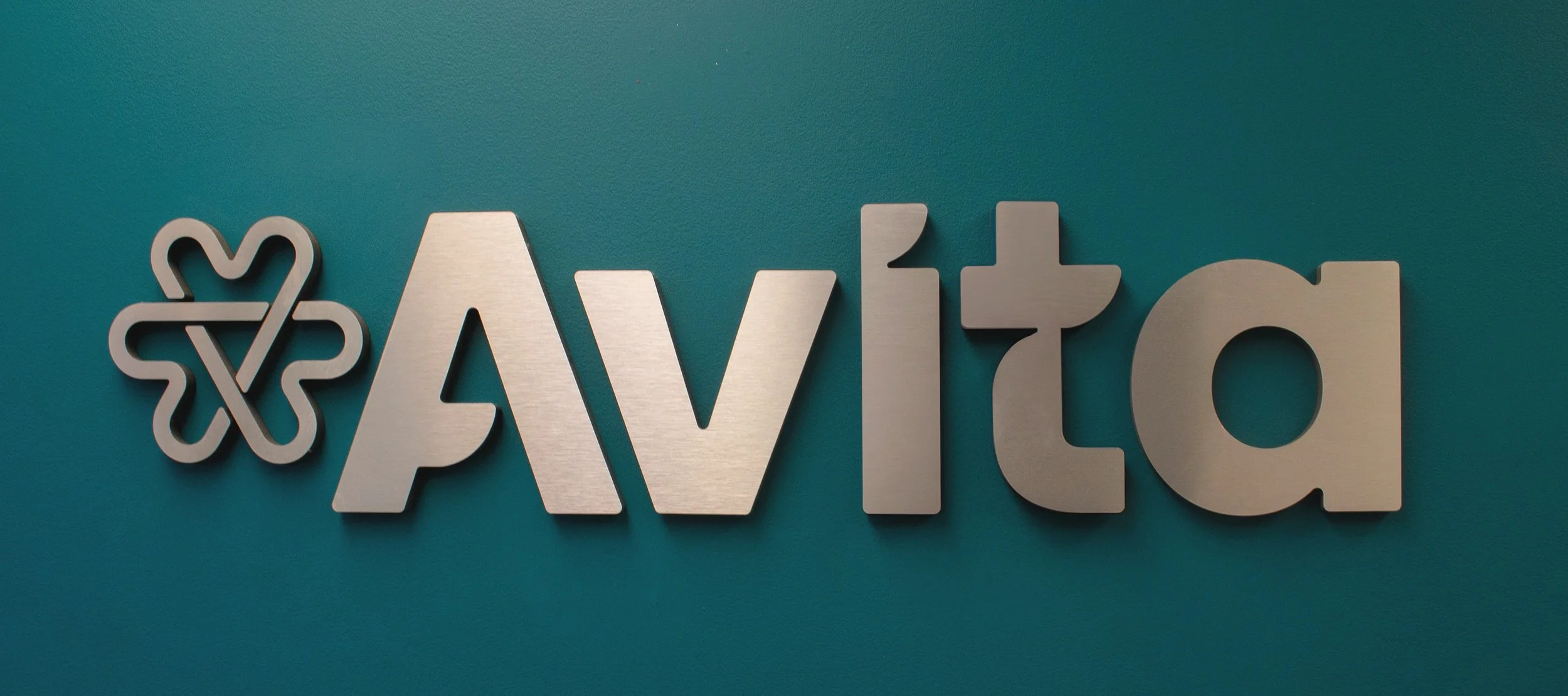 Silver 3D logo of AVITA on teal wall, with a stylized flower-like emblem to the left.