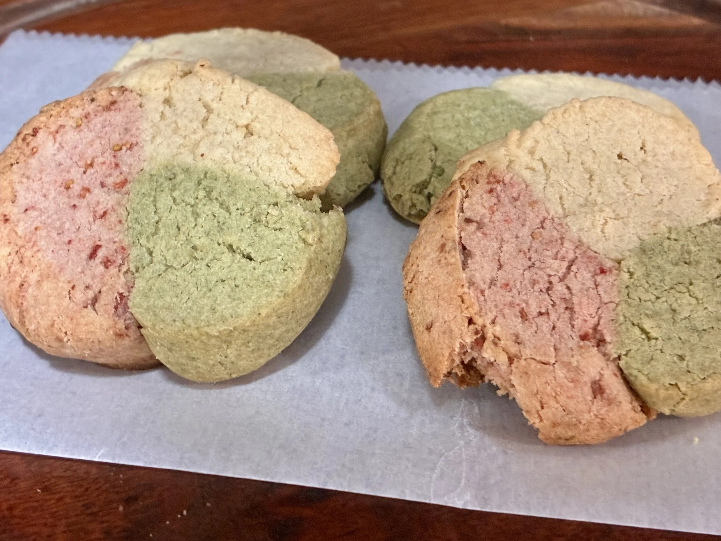 Four cookies sit on a piece of parchment paper. The cookies have three lobes of color, each section's natural flavor lending the cookies their unique appearance. Pink is strawberry, green is matcha, and the usual cookie colored section is vanilla.
