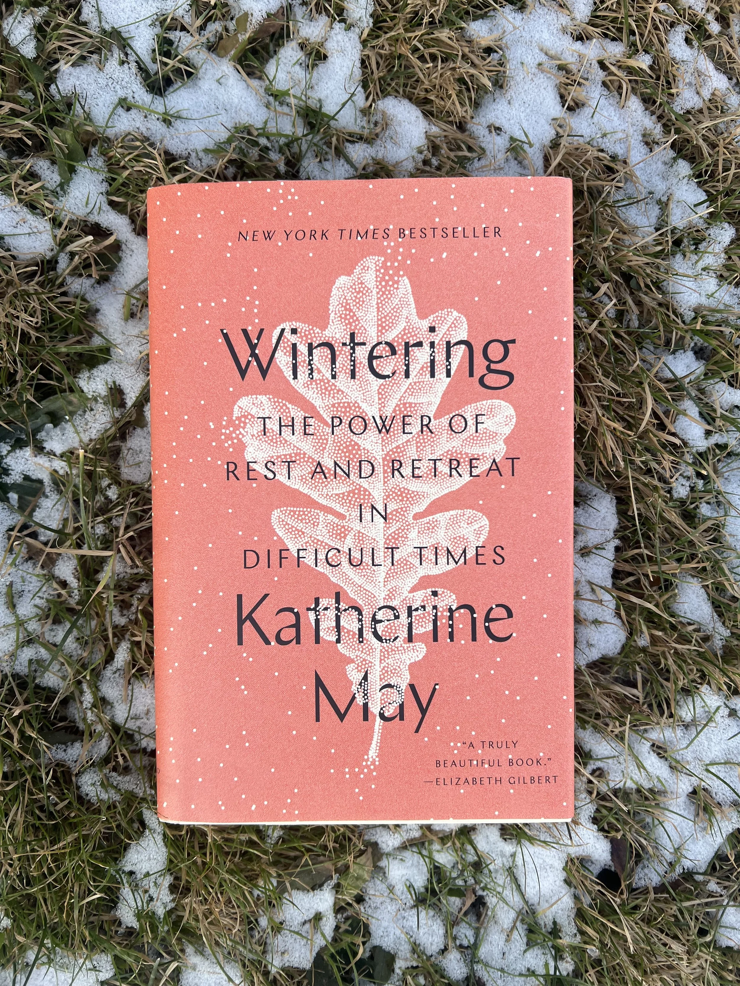 A book rests on green and brown grass poking up through patches of white snow. The orange-pink cover of "Wintering, The Power of Rest and Retreat in Difficult Times" by Katherine May contrasts brightly with its mid-winter resting place.