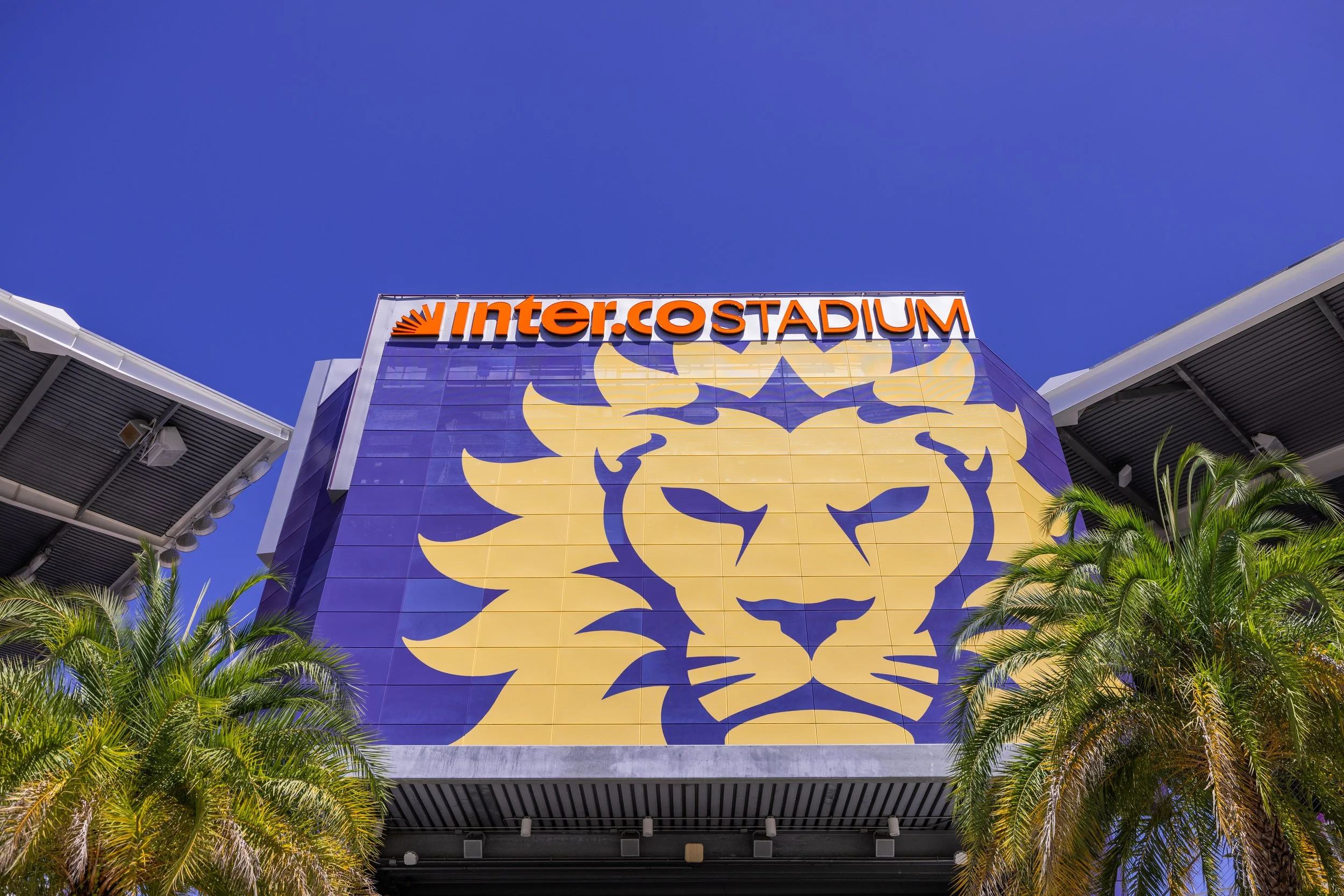 The exterior of the Intuit Dome at Inglewood, California, with a bright yellow and purple lion logo and palm trees in the foreground against a clear blue sky.