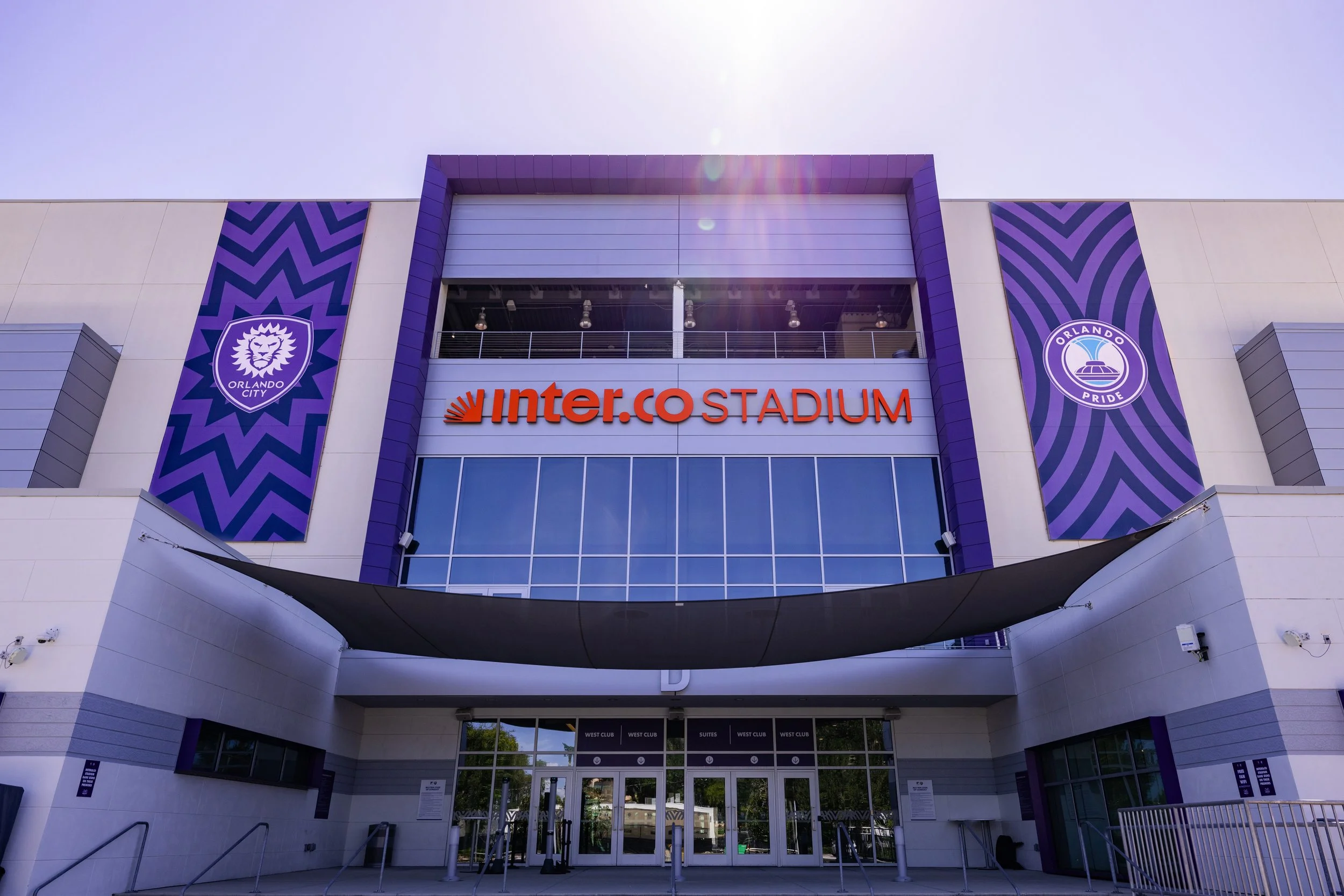 Exterior view of the Orlando City Stadium entrance with purple banners and the 'inter.COSTADIUM' sign, under a clear sky.