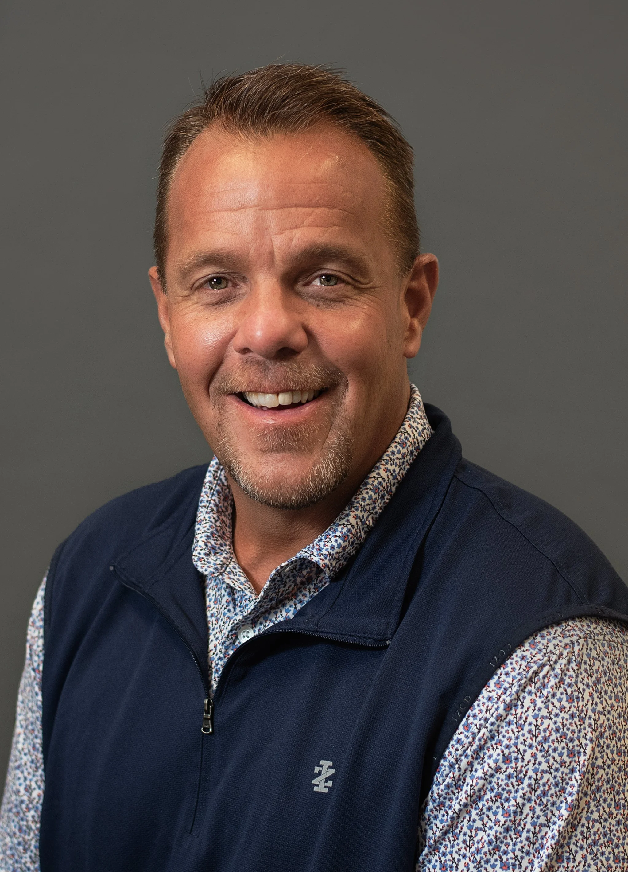 Portrait of Charlie Braam, a middle-aged man with short light brown hair, wearing a navy zip-up vest against a gray background.