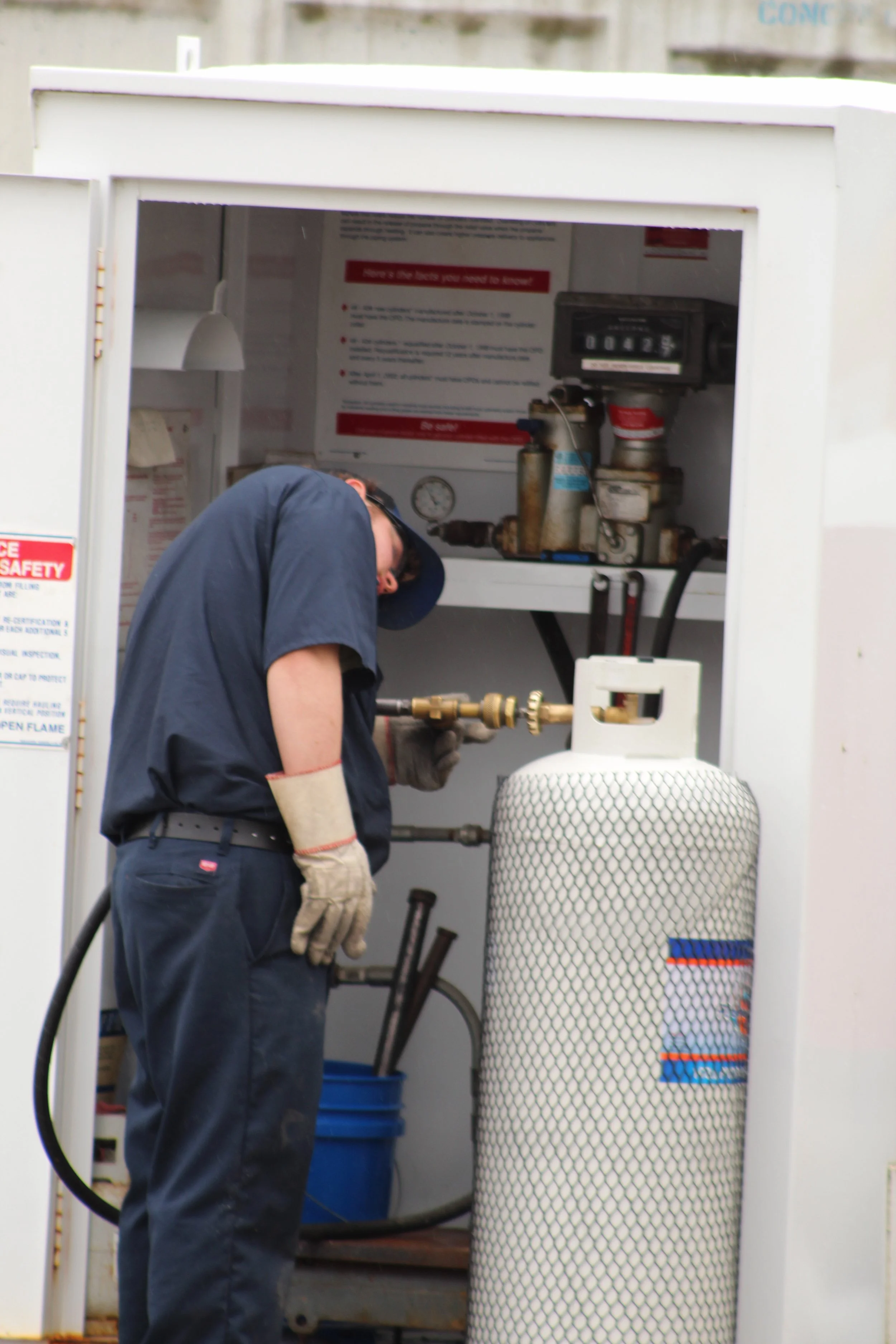 A technician in a navy uniform and gloves working with a gas tank and machinery inside a safety enclosure, with tools and informational signs visible in the background.