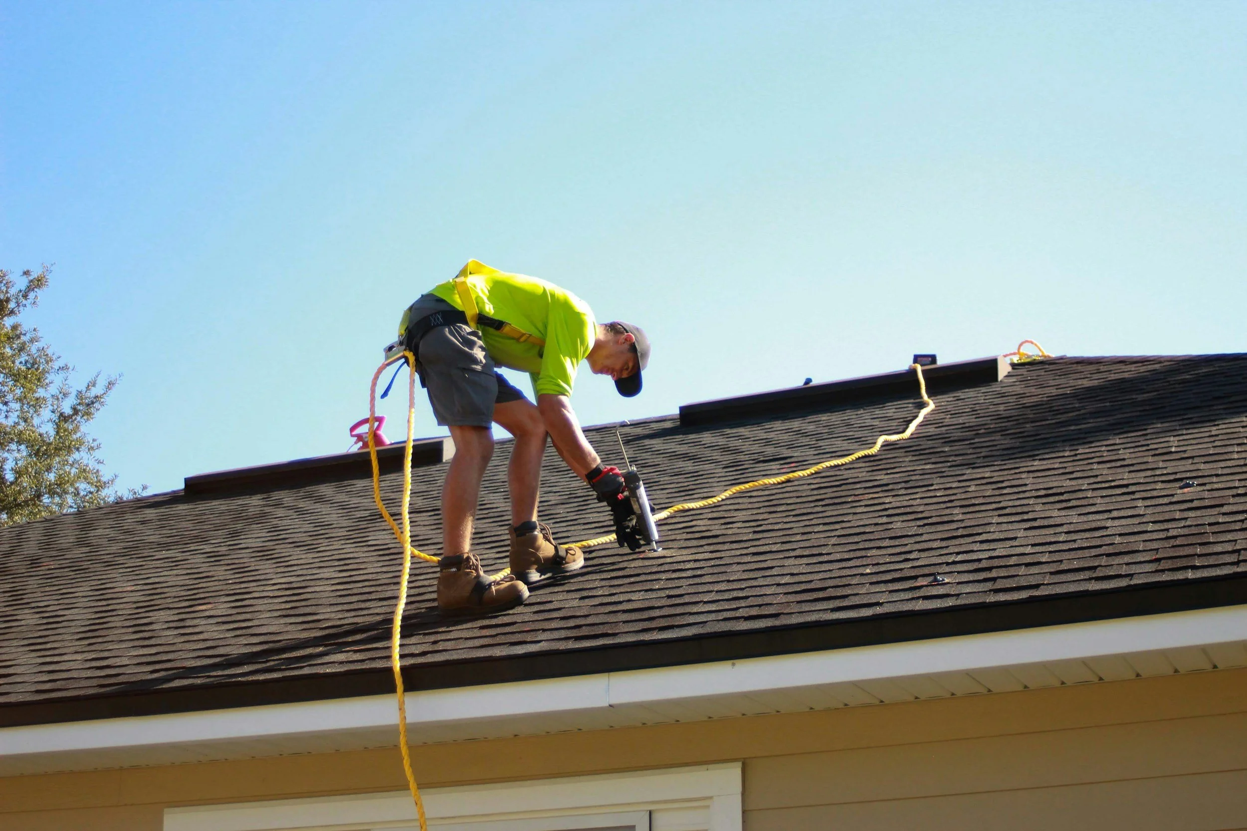 A man in a yellow shirt, shorts, and work boots is on a rooftop, working with a power tool, secured with safety harness and ropes, against a clear blue sky.