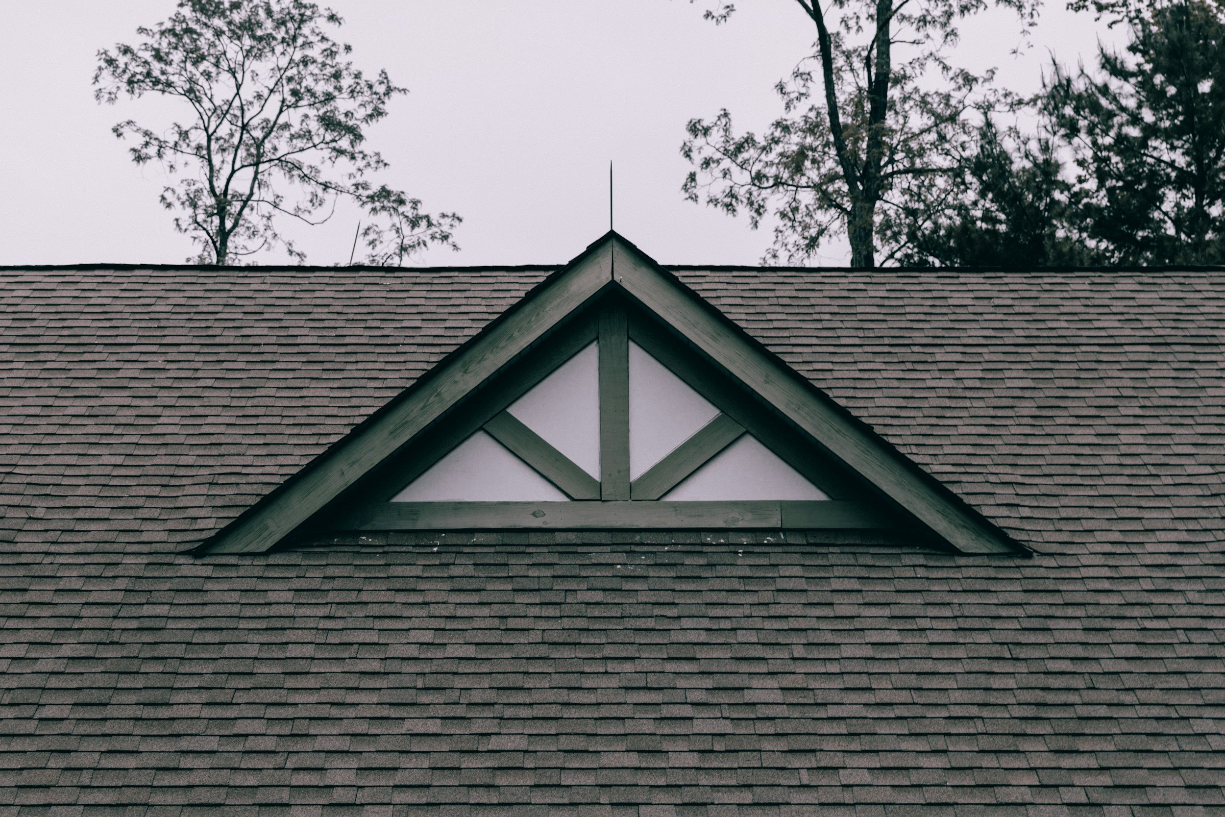 Close-up of a house's gable roof with brown shingles and decorative white trim, with tall trees and an overcast sky in the background.