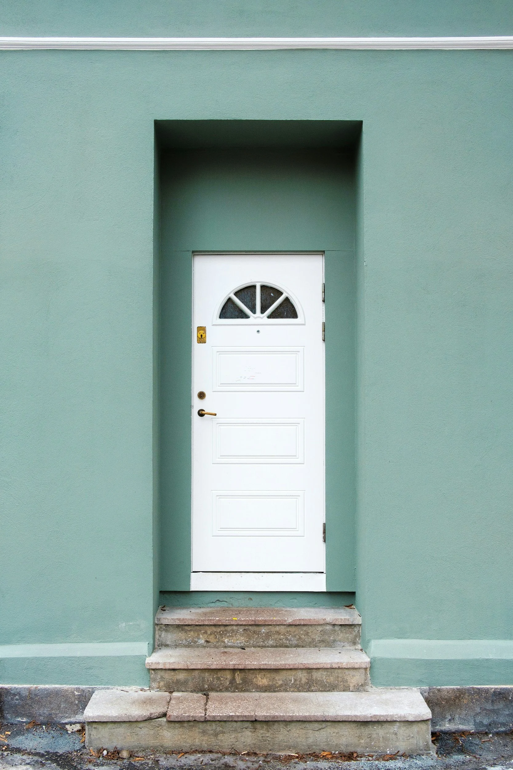 Green building wall with a white front door framed in an alcove, accessible by a short set of three concrete steps.