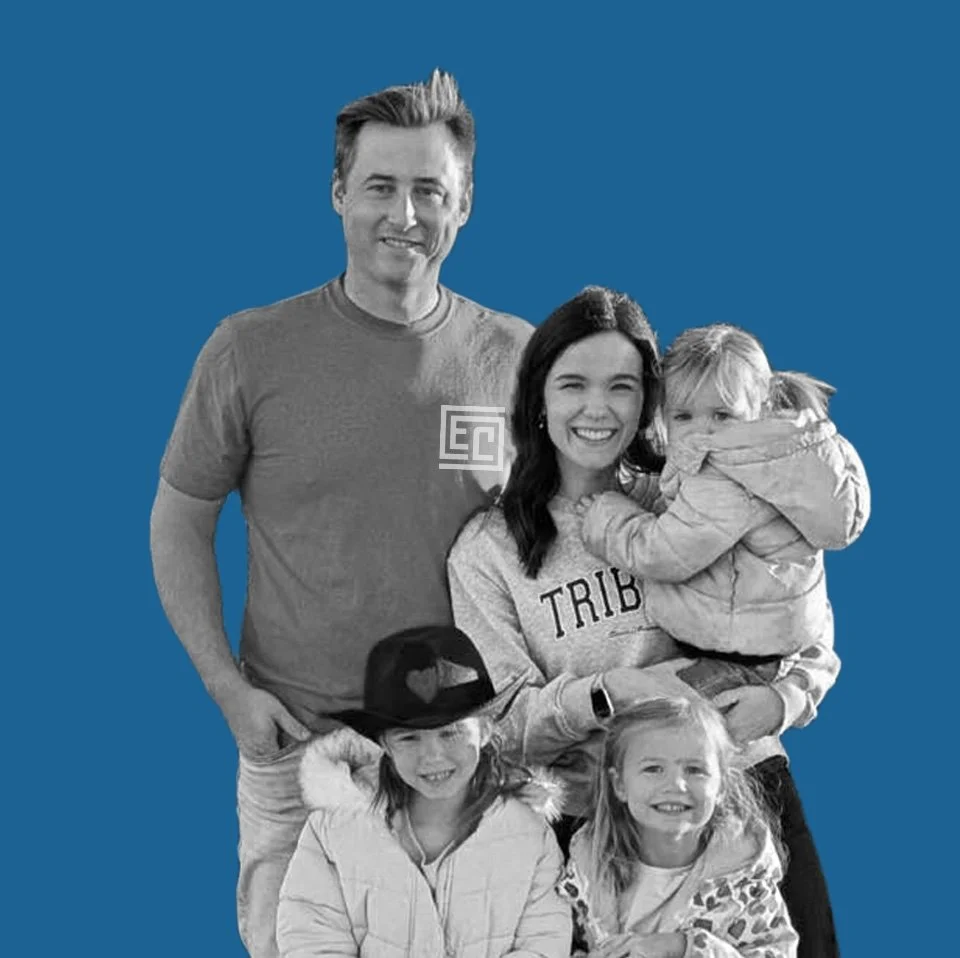 A family of five adults and children posing for a photo against a blue background. One woman is holding a small child, and two young girls are in front of the group, all smiling.