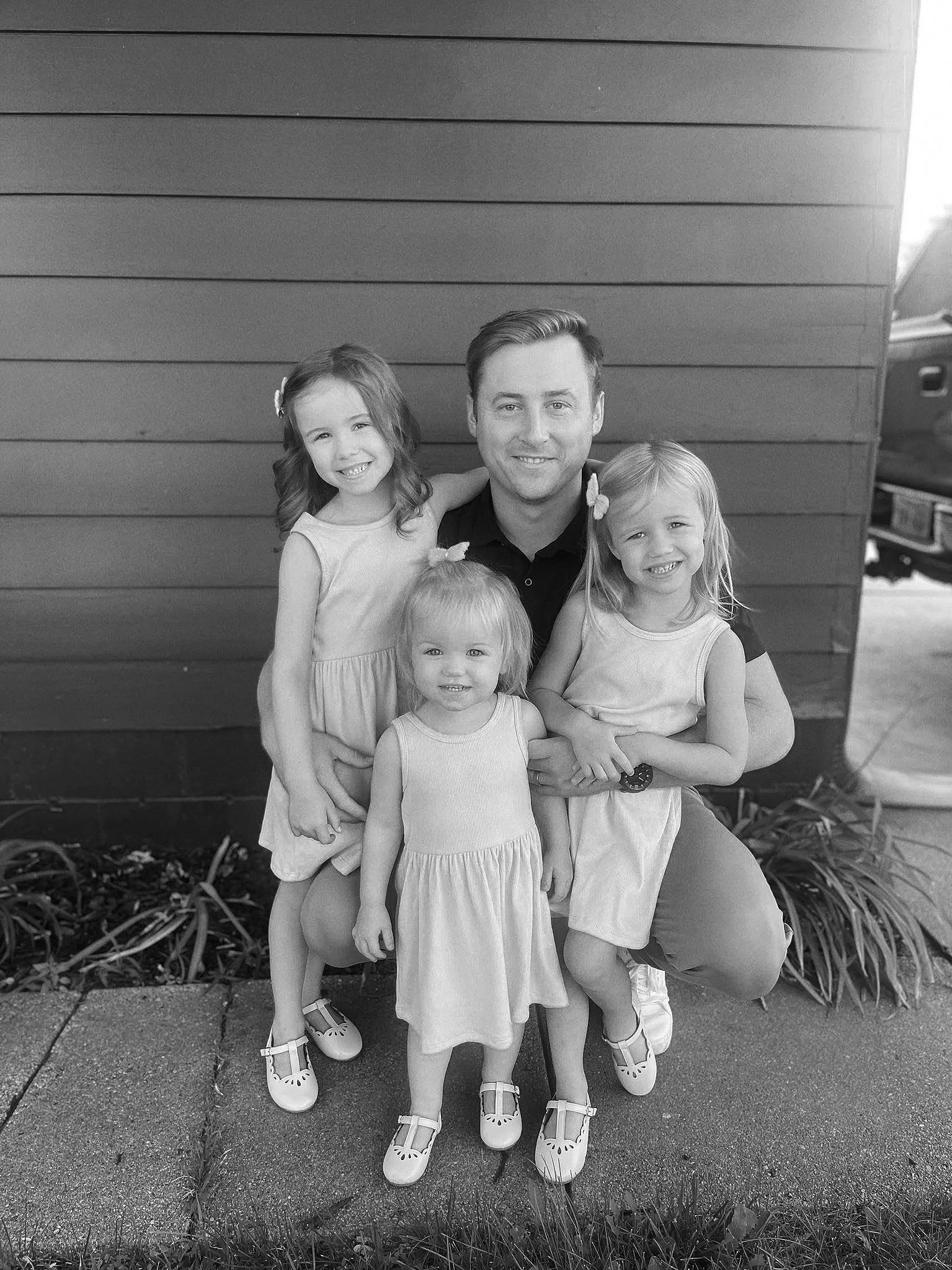 A man with three young girls outdoors in front of a wooden wall, all smiling. They are dressed in light-colored dresses and wearing matching sandals.