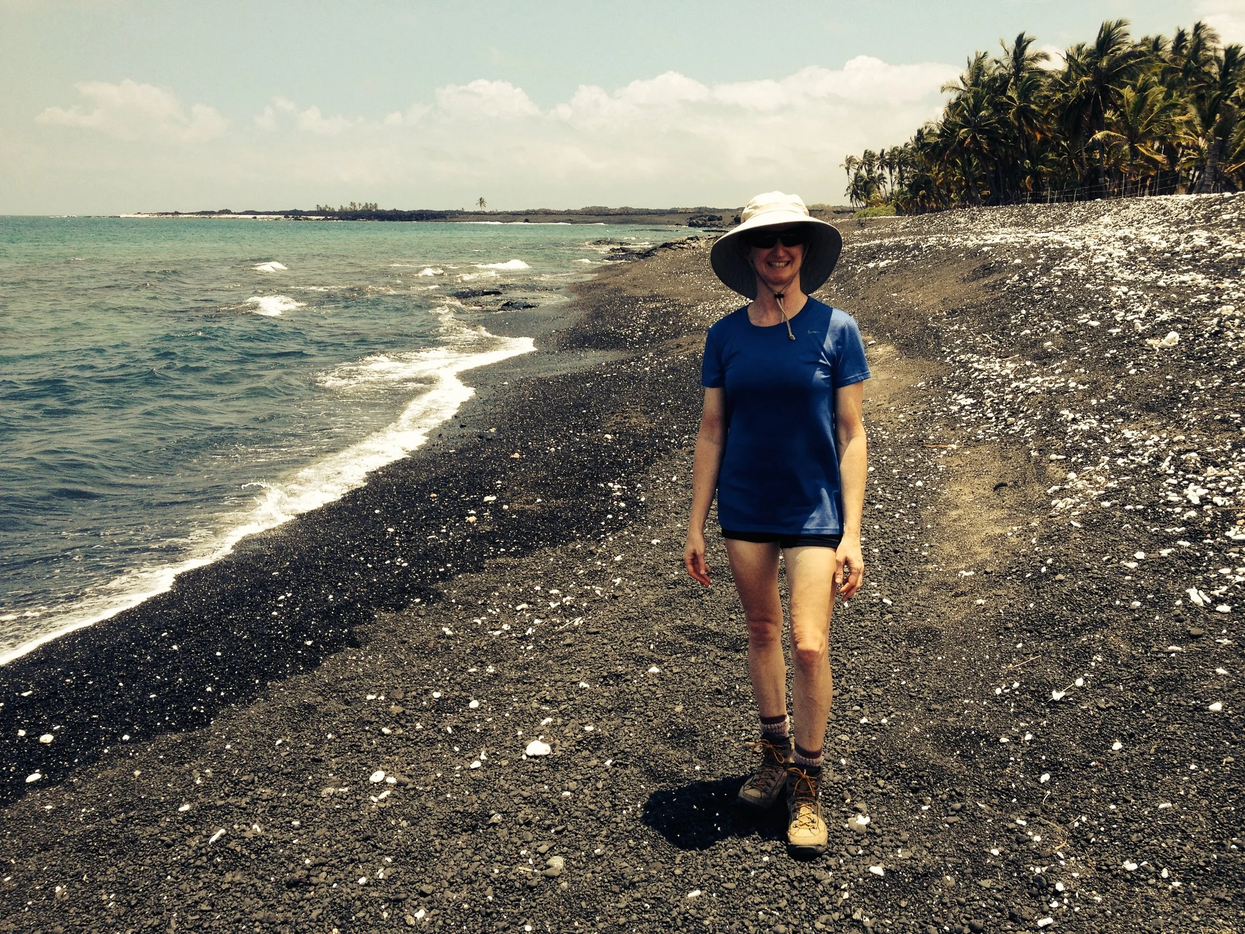 Woman wearing a hat, sunglasses, a blue t-shirt, shorts, and hiking boots walking on a rocky beach near the ocean with palm trees in the background.
