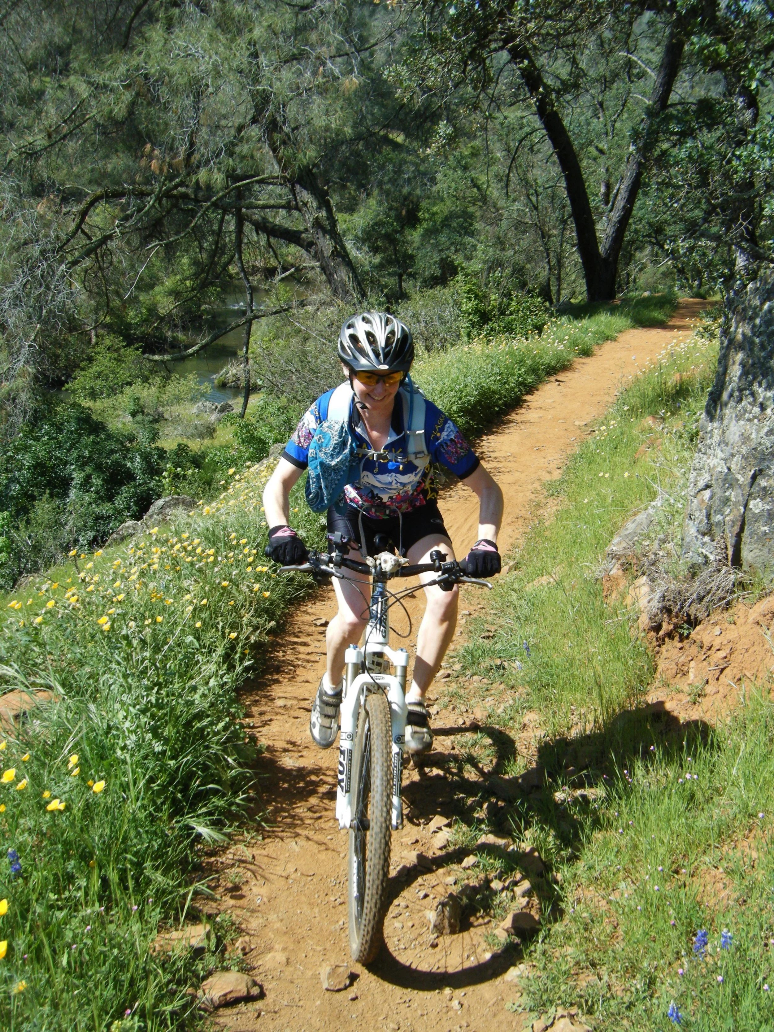 A woman in a helmet and sunglasses riding a mountain bike on a dirt trail surrounded by green trees and wildflowers on a sunny day.