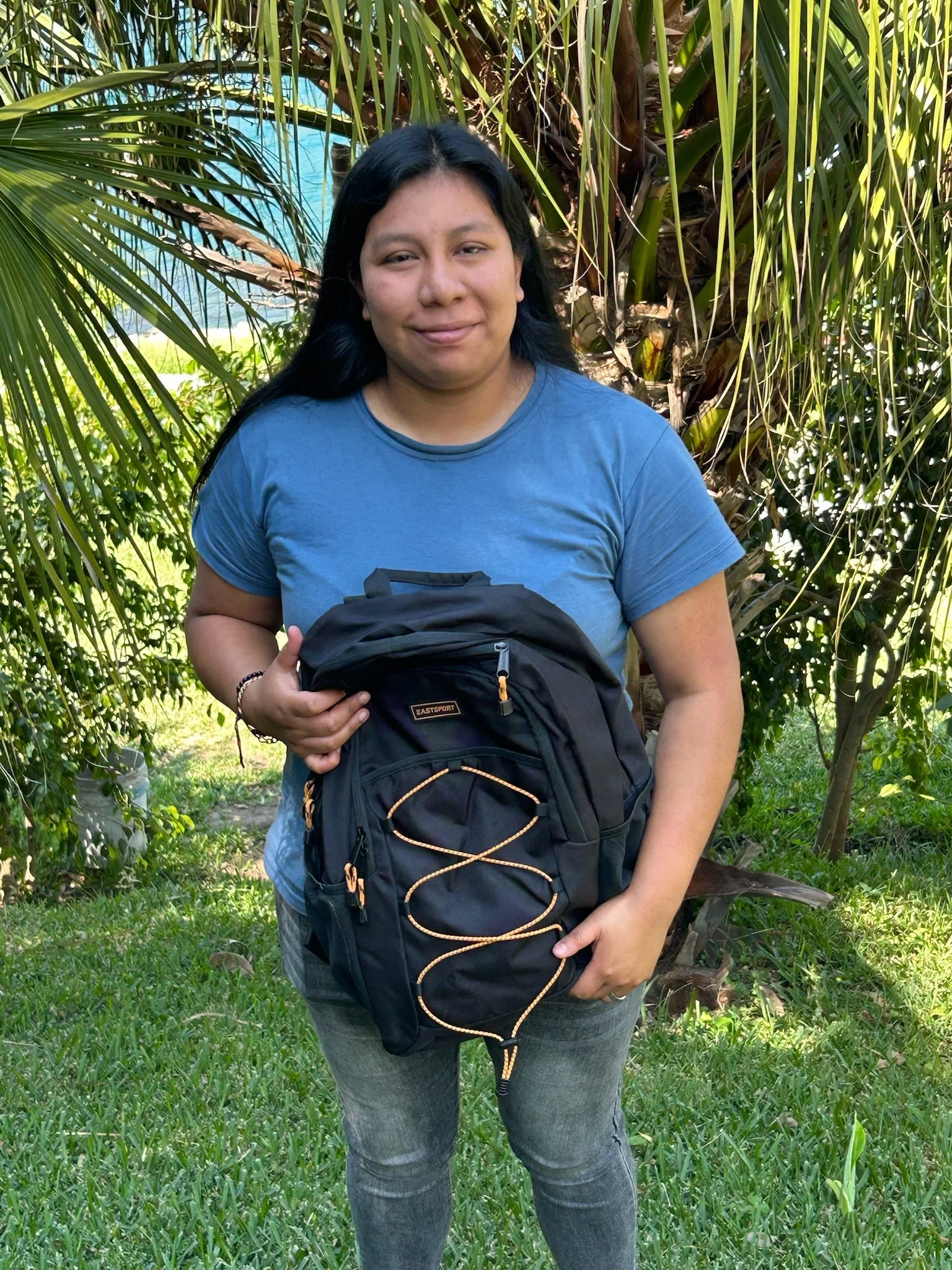 Young woman standing outdoors in front of tropical plants, wearing a graphic t-shirt, jeans, and sneakers, smiling at the camera.