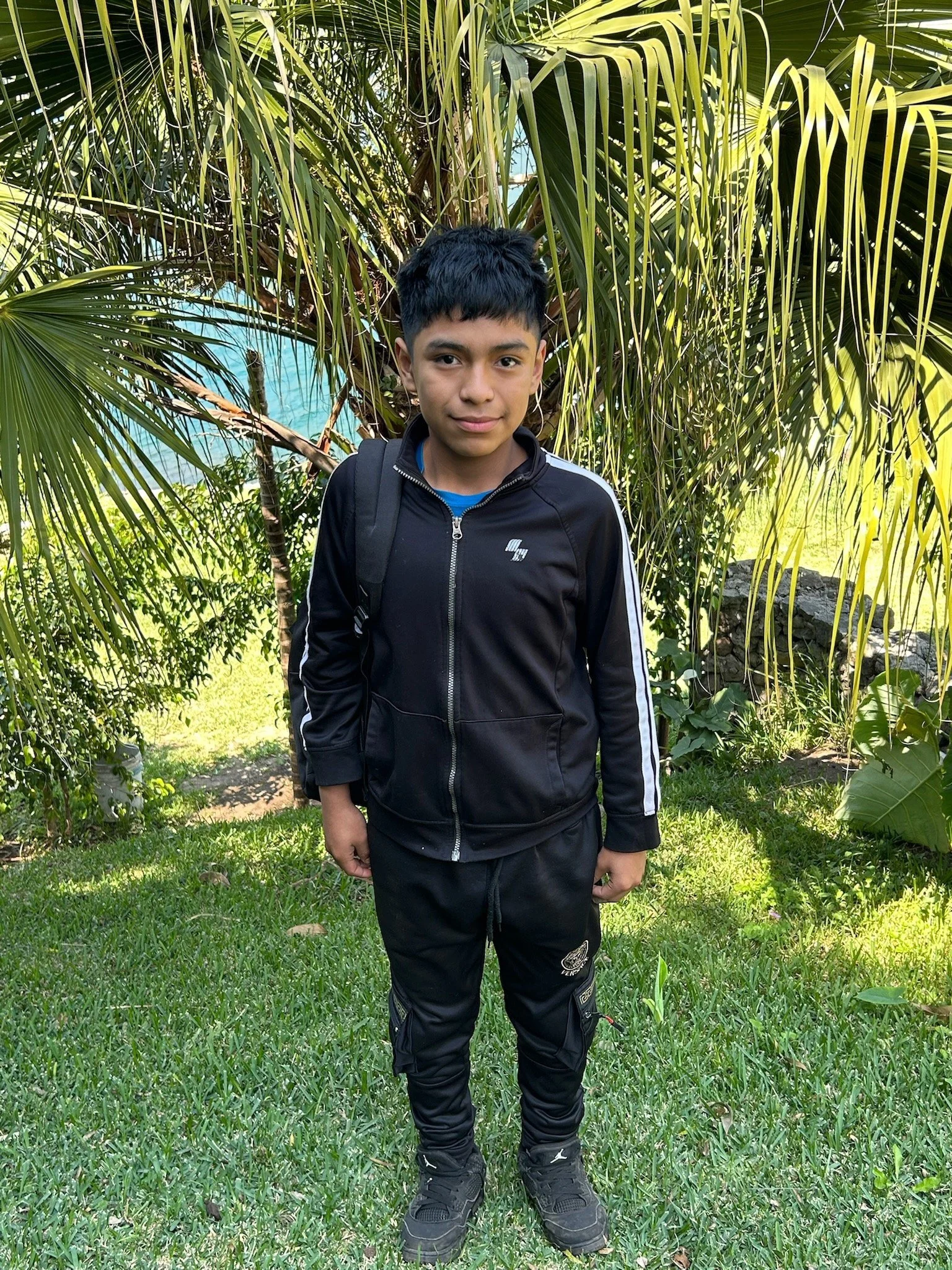 A young boy standing outdoors with a volcano in the background. He is wearing a black and navy Sonic the Hedgehog sweatshirt and has short dark hair.