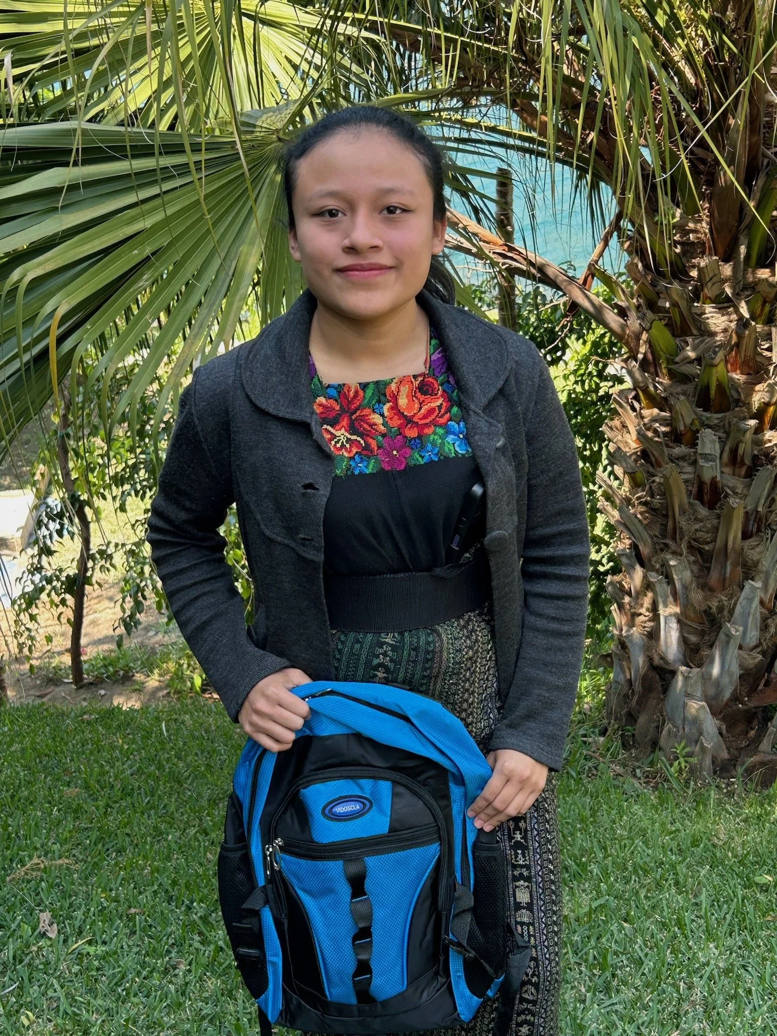 A young woman in traditional Guatemalan clothing standing outdoors with a volcanic mountain in the background and blue sky with clouds.