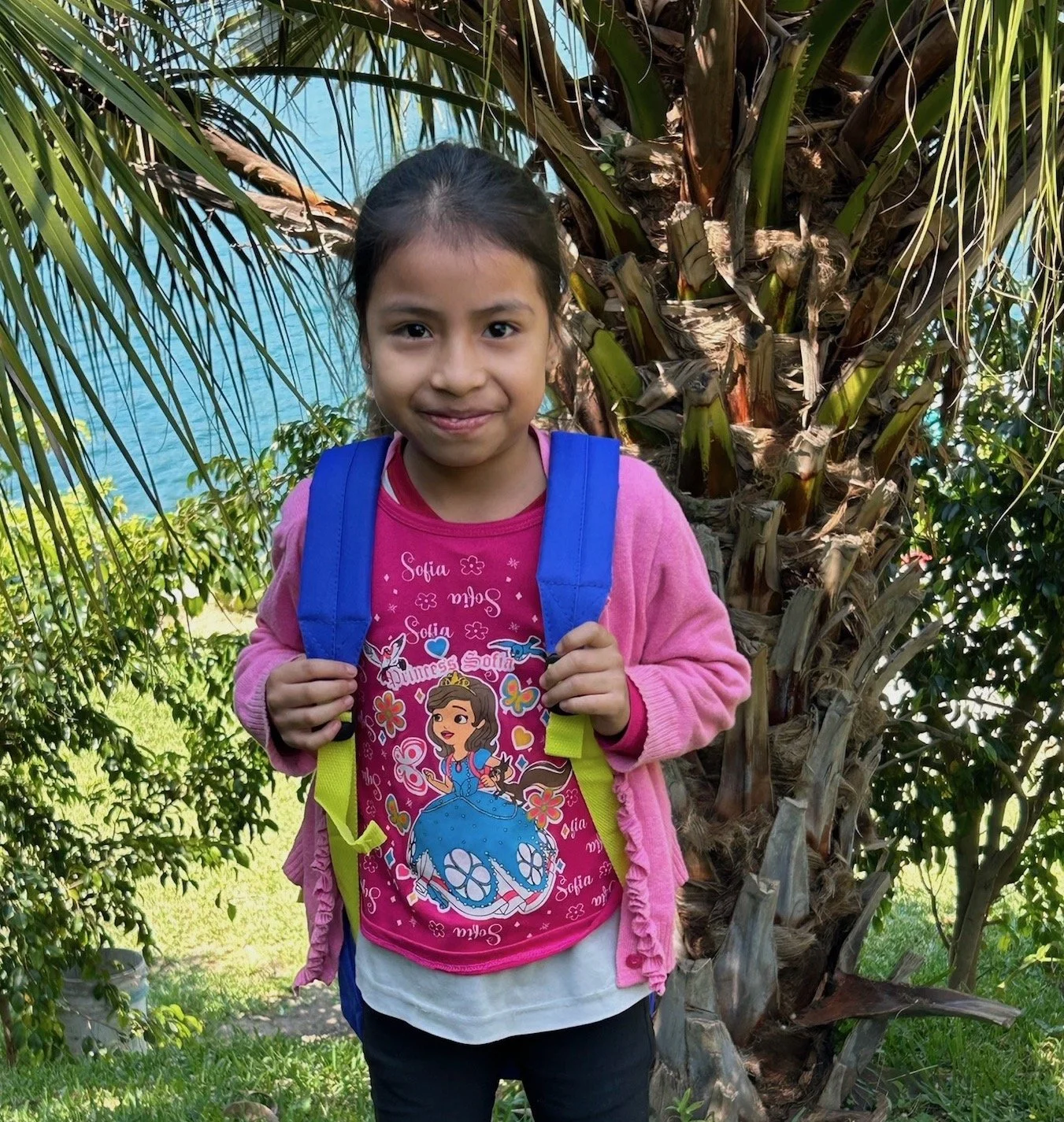 A young girl wearing an orange Snoopy t-shirt standing outdoors with a mountain and volcano in the background, under a blue sky with clouds.