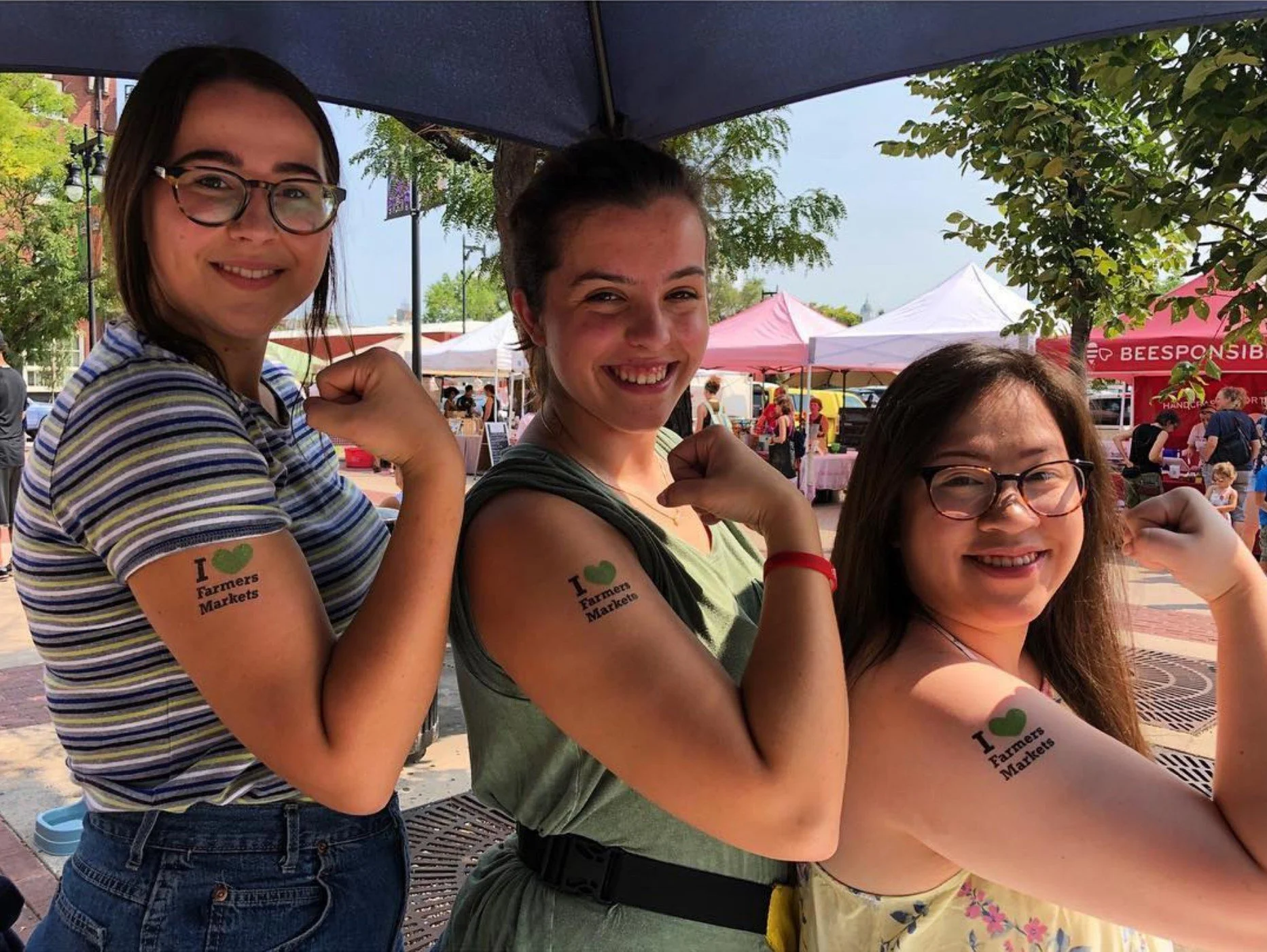 Three women at an outdoor event, each flexing their right arm with temporary tattoos on their upper arms that read "I ♥ Farmers Markets." They are smiling and under a canopy with tents and people in the background.