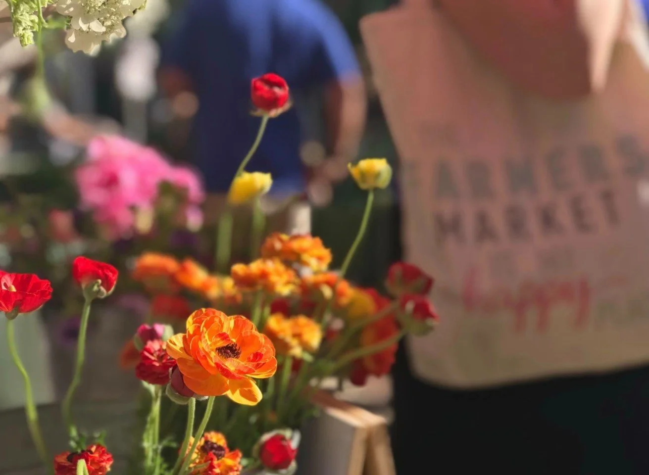 Close-up of colorful flowers, including orange, yellow, and red blooms, at a market stall with blurred people in the background.