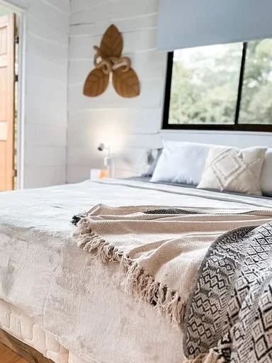A cozy bedroom with a large bed covered in a beige blanket and decorative pillows, a window overlooking trees, a nightstand with a lamp, and a wall decoration of a moth.