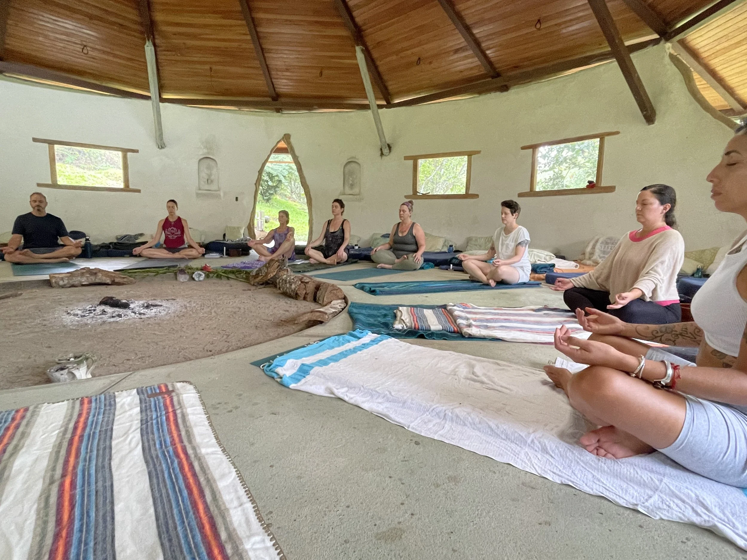 People participating in a meditation or yoga session seated in a circle inside a rustic room with wooden ceiling and small windows, with a campfire ring in the center.