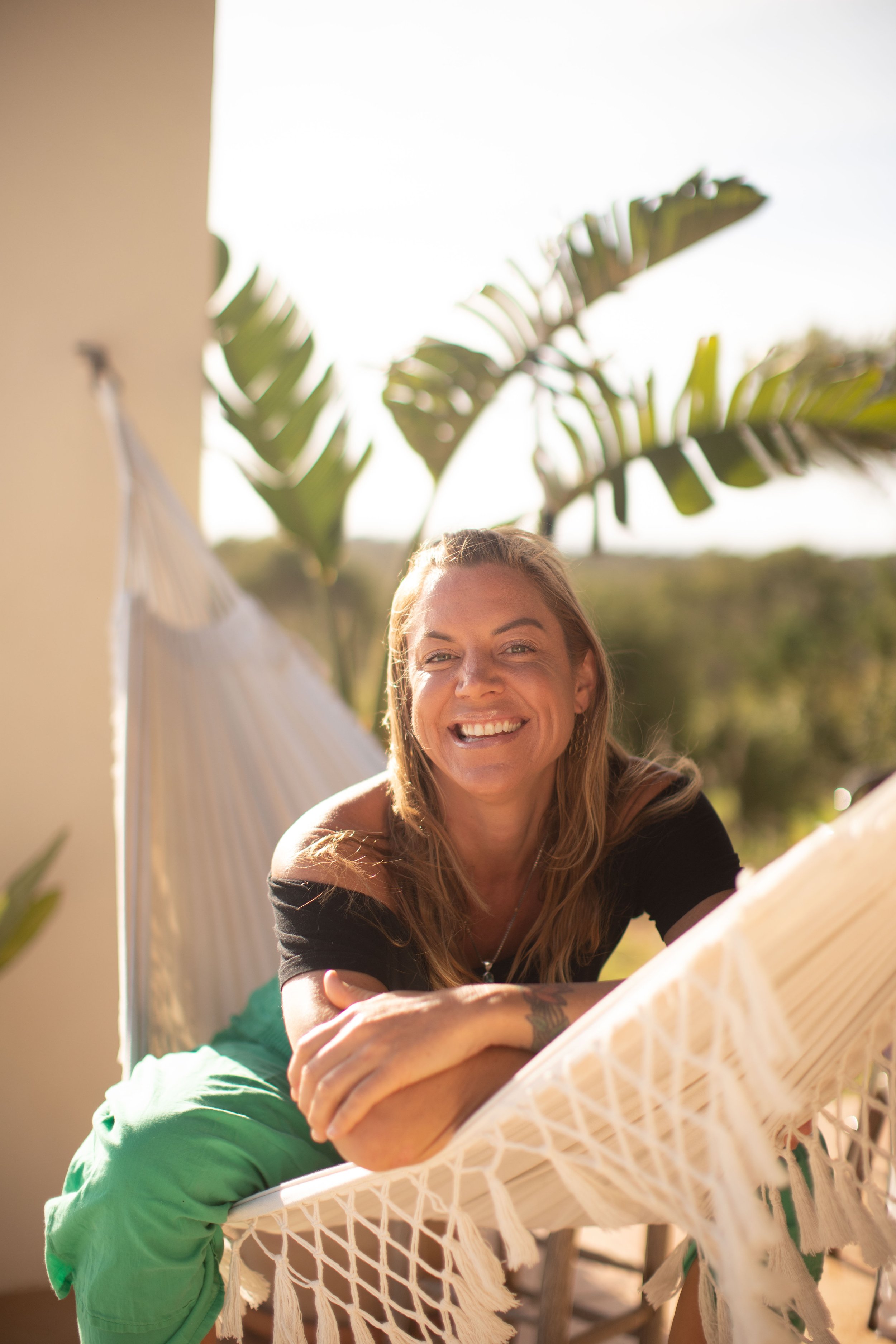 A smiling woman with blonde hair relaxing outdoors on a white hammock with green leaves in the background.