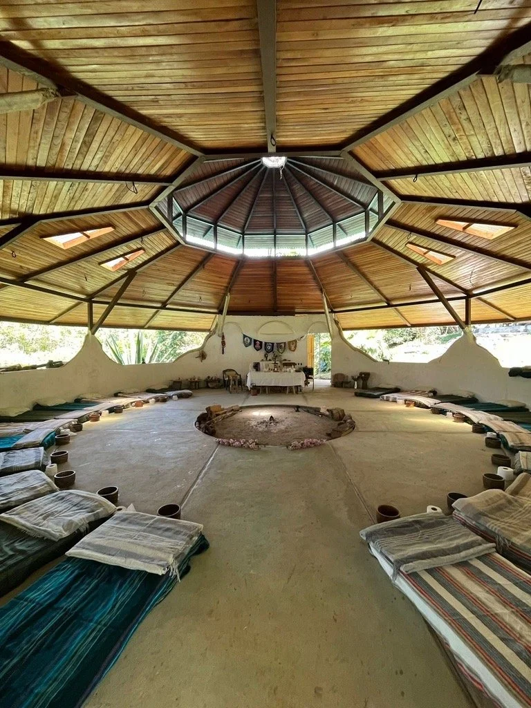 Circular indoor space with a wooden roof, cushions and small tables arranged along the perimeter, and a central fire pit decorated with flowers. There is a table with items and banners or flags on the wall at the far end.