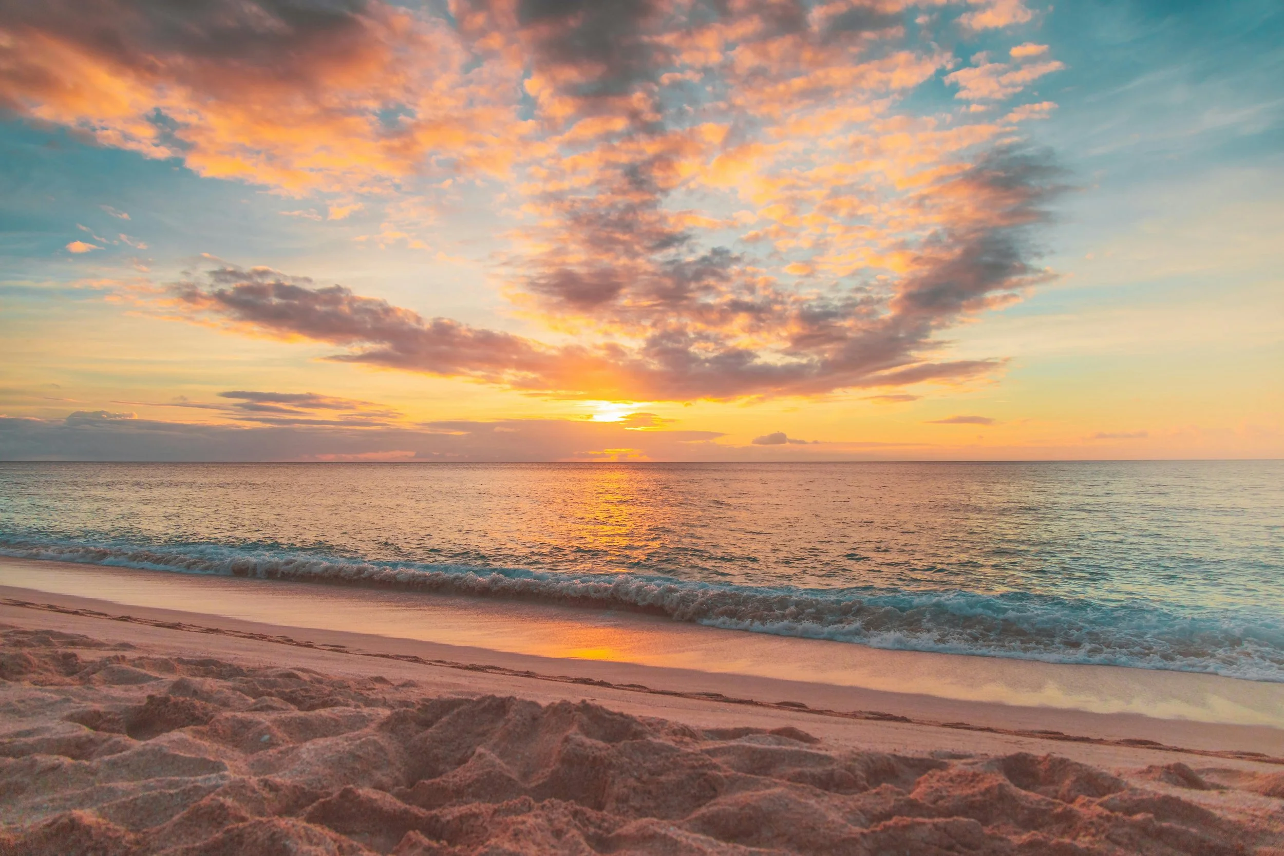 Sunset over the ocean with colorful clouds, calm waves, and sandy beach in the foreground.