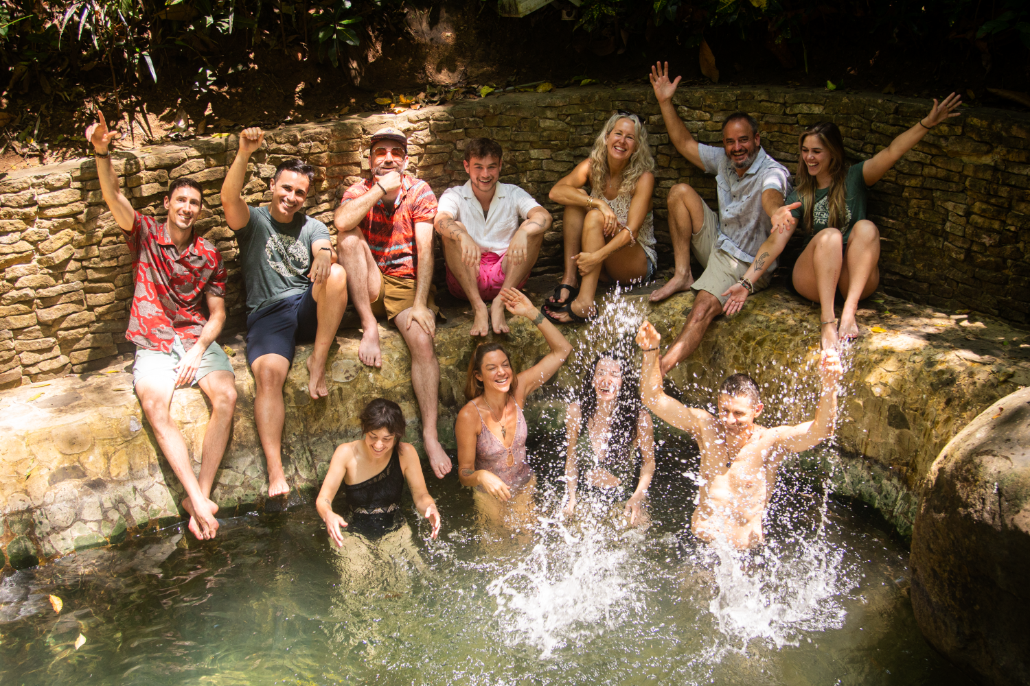 A group of people enjoying themselves in a small pool with a stone and brick wall background, some sitting on the edge and others splashing in the water.