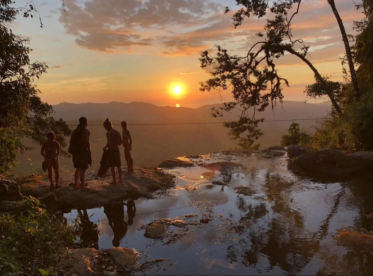 Silhouetted group of five people standing on rocks near a waterfall at sunset, with trees and mountains in the background.